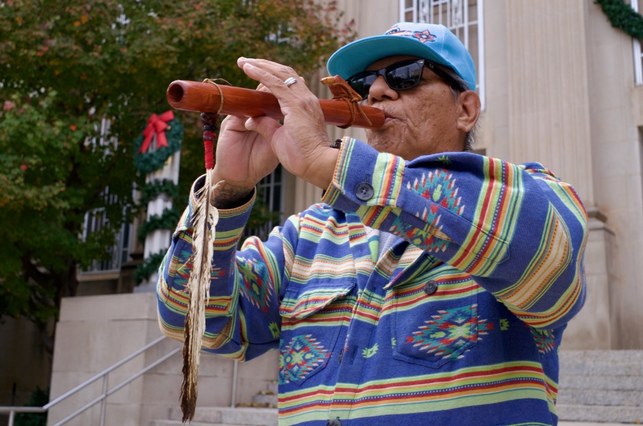 An elderly man wearing a blue cap, sunglasses, and a colorful patterned jacket playing a wooden flute outdoors.