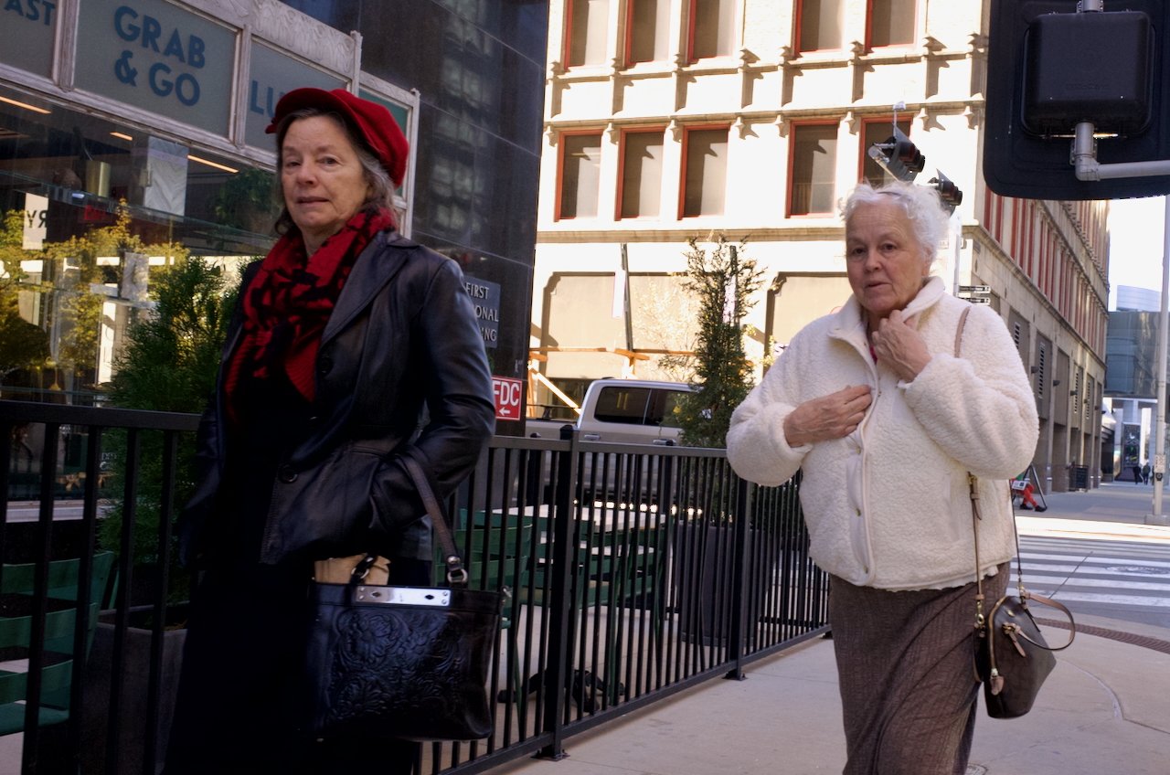 Two elderly women walking on a city street, one in a black leather jacket with a red scarf and hat, the other in a white fleece jacket with a purse, near a crosswalk and buildings.