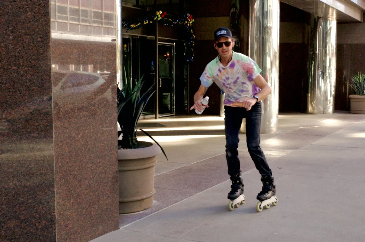 A man wearing sunglasses, a colorful tie-dye shirt, and a cap skateboarding on a city sidewalk, holding a water bottle.