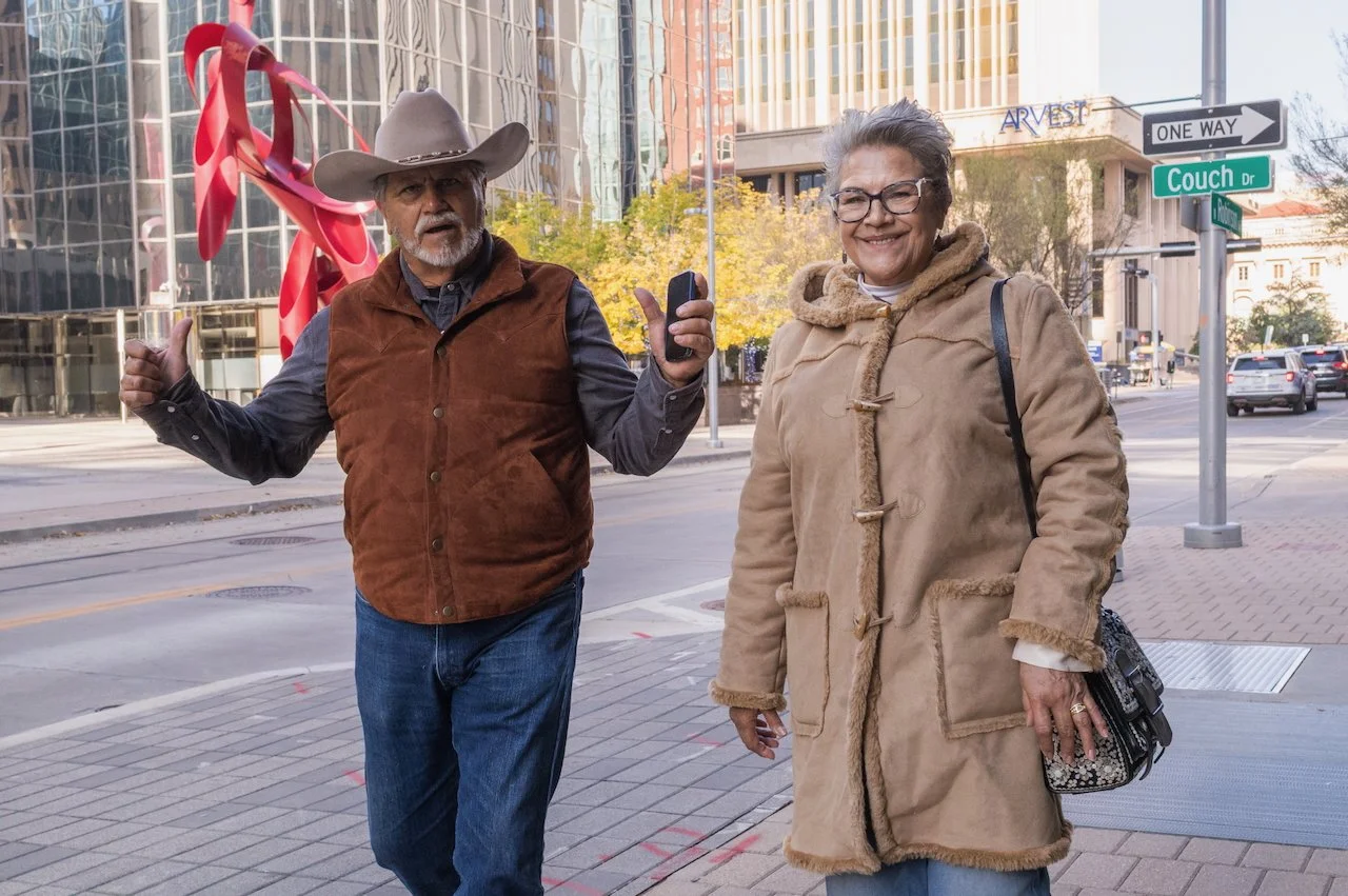 A man wearing a cowboy hat, brown vest, and jeans giving a thumbs up, and a smiling woman in a tan coat and glasses standing on a city sidewalk with buildings and street signs in the background.
