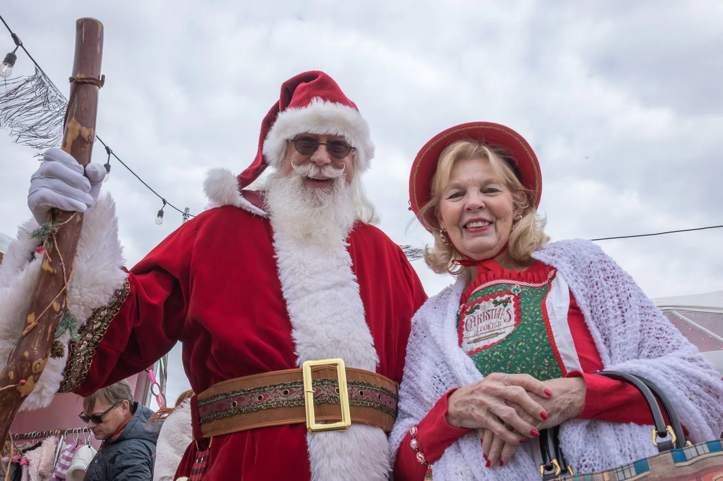 A man dressed as Santa Claus and a woman dressed in Christmas attire smiling at the camera during a holiday event.