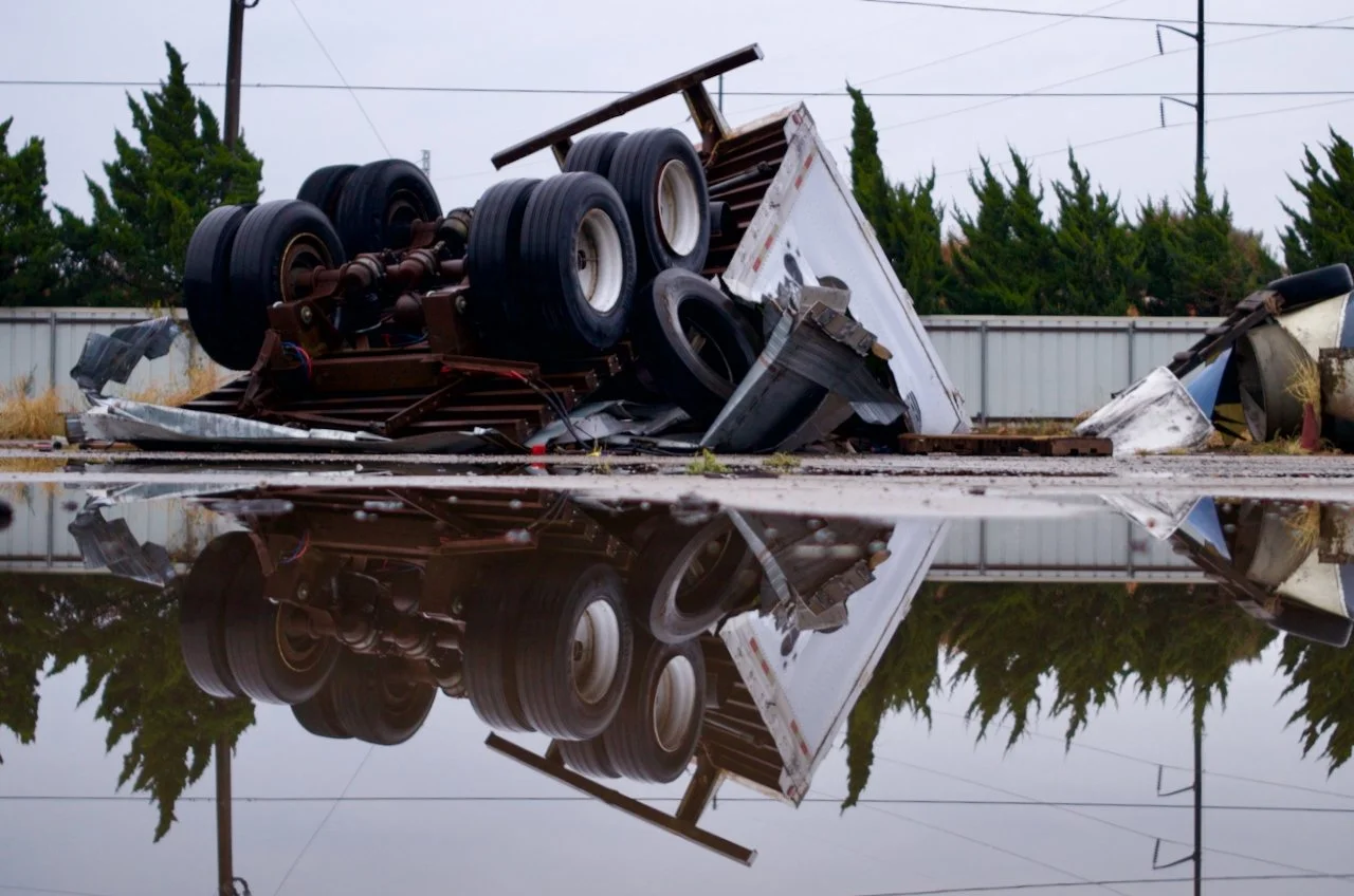 Upside-down overturned truck and debris in a puddle reflecting the scene on a cloudy day.