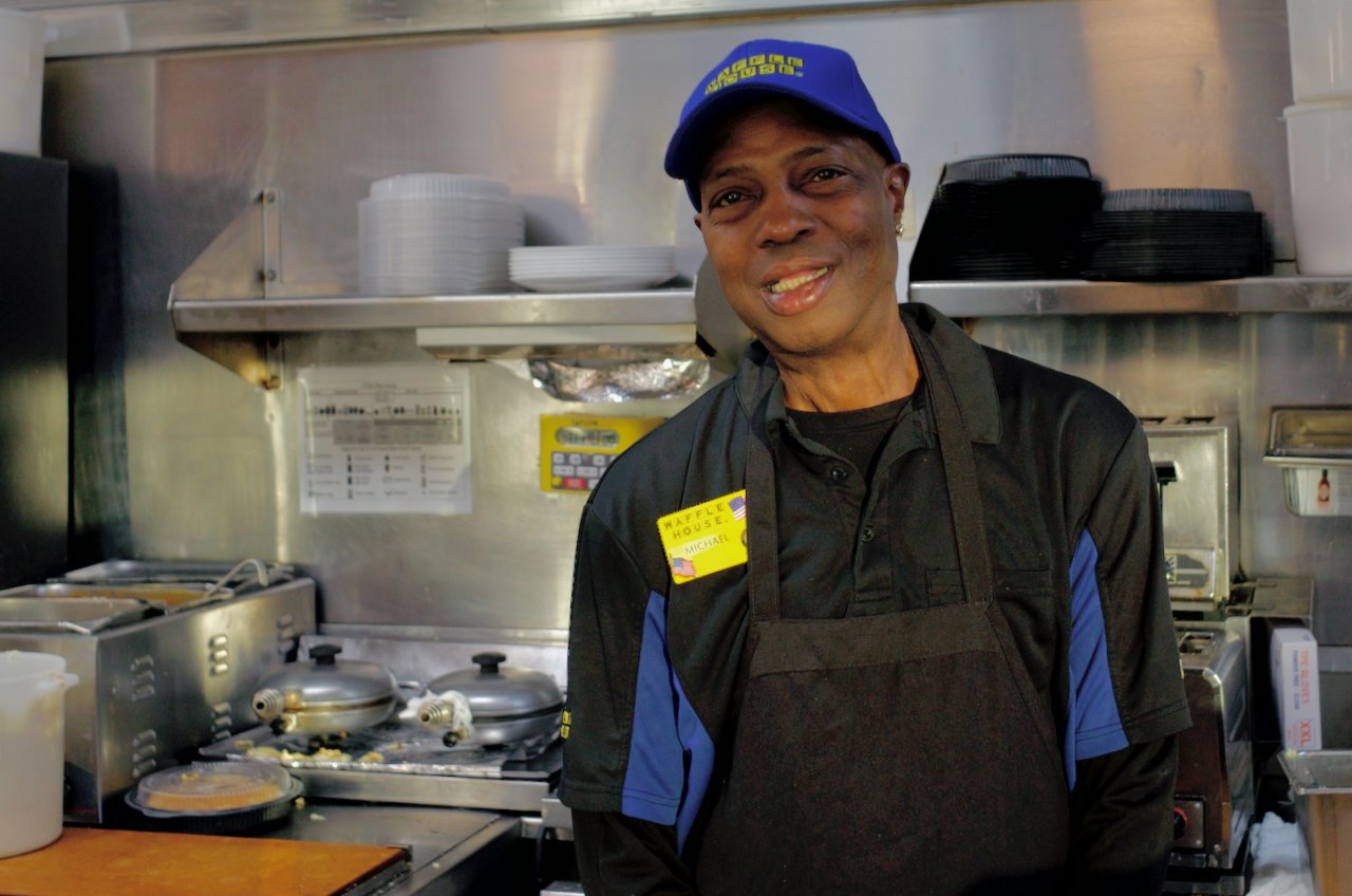 A smiling woman wearing a blue cap, black apron, and black shirt standing in a commercial kitchen with kitchen equipment and dishes in the background.