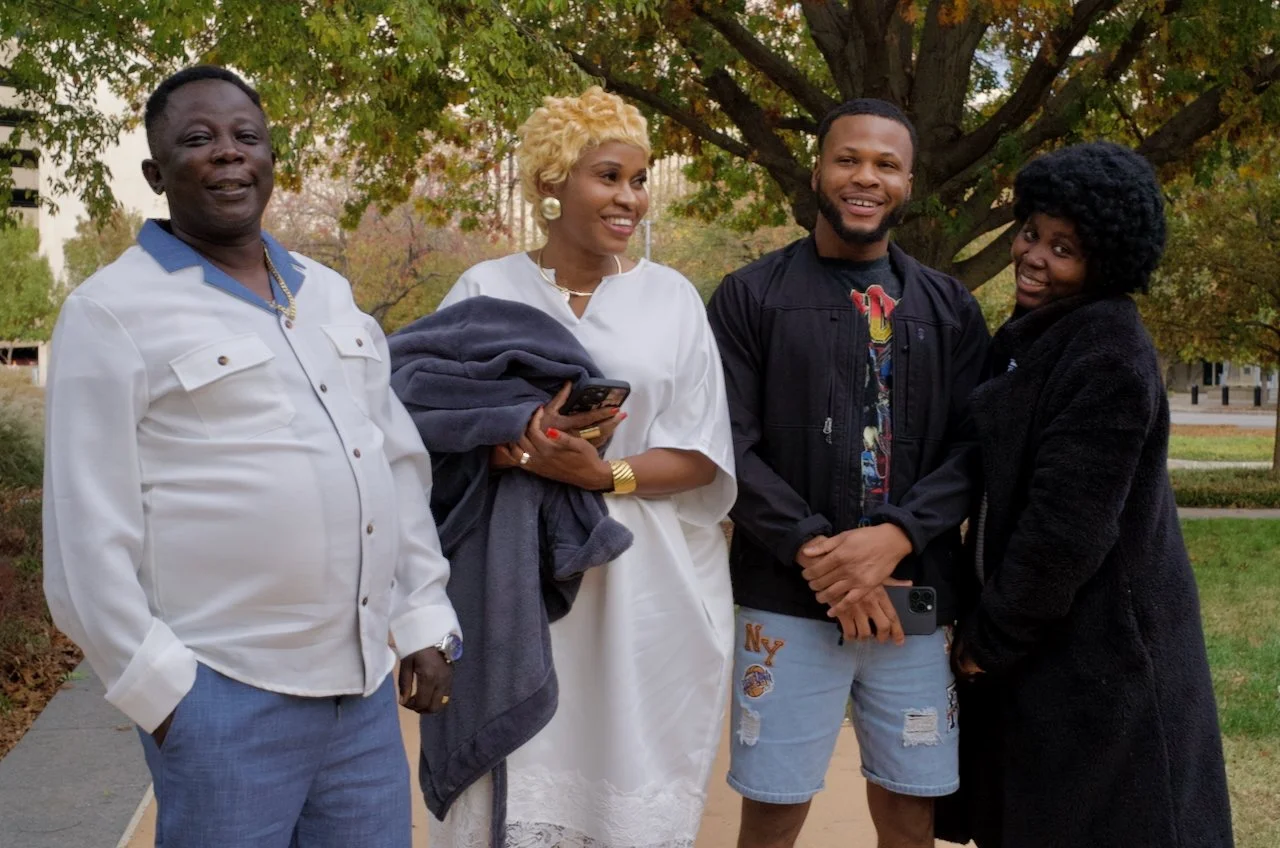 Four people stand outdoors under a tree, smiling and chatting. From left to right: an older man in a white shirt and blue pants, a woman with short blonde hair in a white dress holding a jacket and phone, a young man in a black jacket and shorts, and
