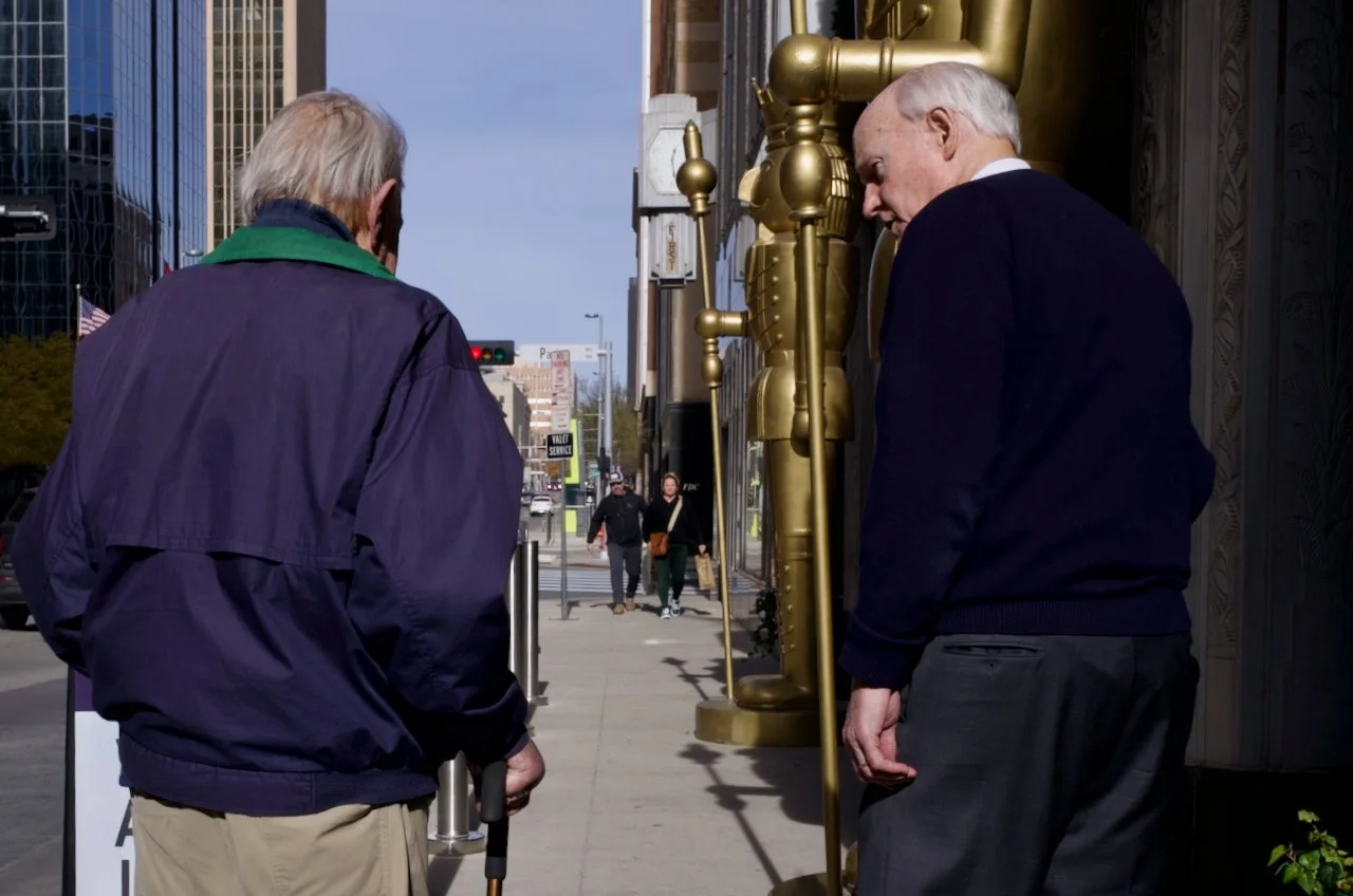Two elderly men standing outside a building, engaged in conversation. One man is facing away, holding a cane, wearing a dark jacket with a green collar and beige pants. The other man is facing the building, dressed in a black sweater and gray pants. 