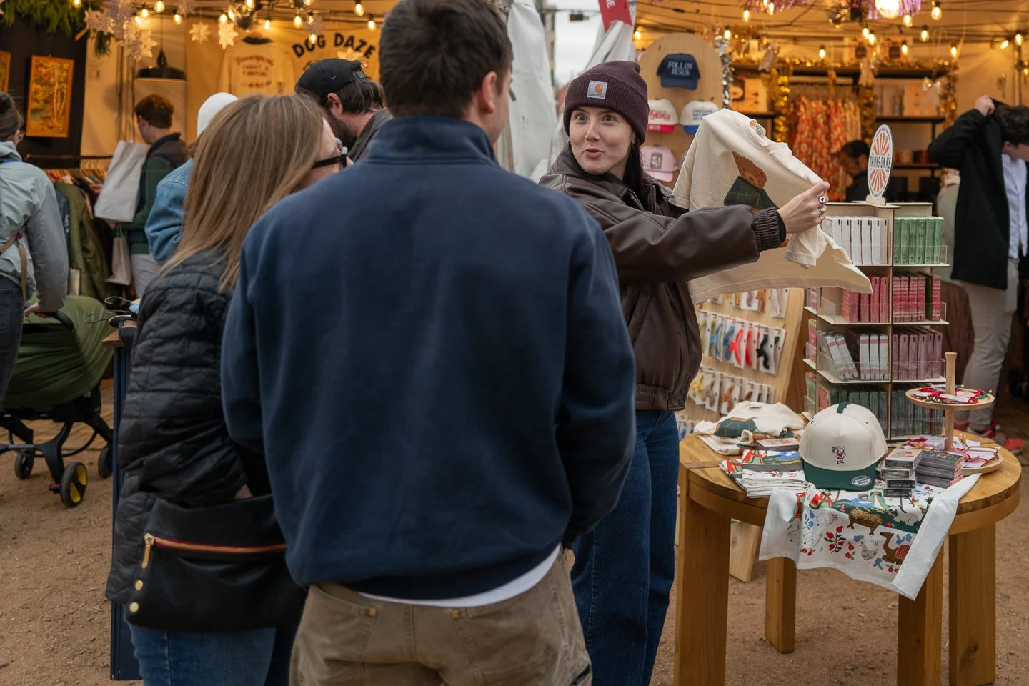 A woman at a market stall distributing a printed cloth to customers, with a display of gifts including hats, small boxes, and Christmas-themed items on a wooden table, and other shoppers browsing in the background.