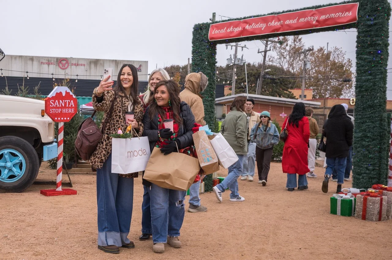People taking a selfie at a holiday market decorated with Christmas lights and ornaments. Gift boxes are on the ground nearby, and a red sign says 'Santa Stop Here'. The holiday arch has a sign that reads 'Local shops, holiday cheer and merry memorie