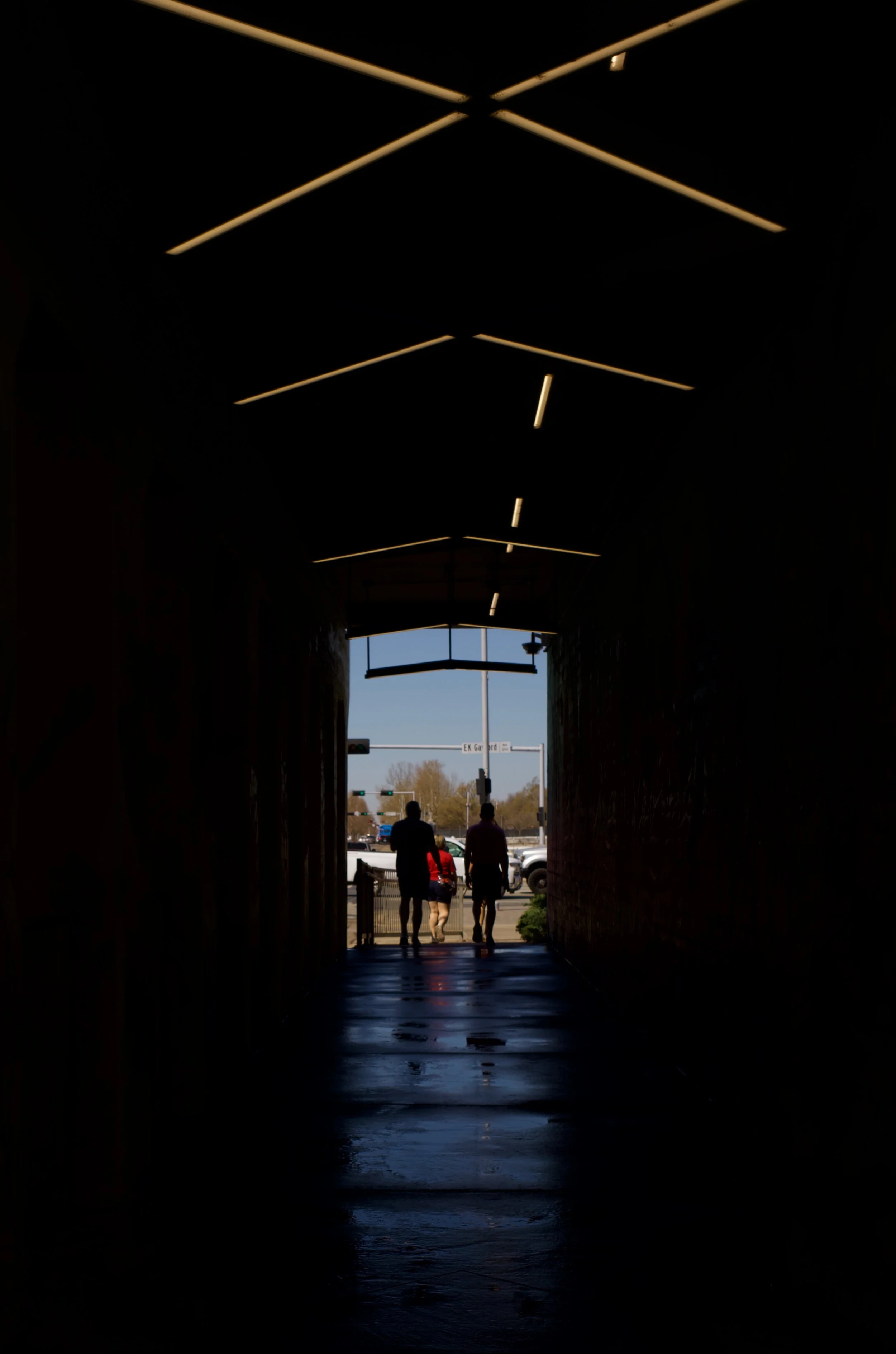 Silhouette of three people walking through a dark tunnel towards a sunny street with traffic lights and cars.