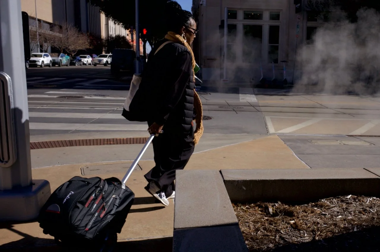 A woman with glasses, a black jacket, and a tan scarf pulls a rolling backpack while walking on a city sidewalk with steam rising from a vent nearby.