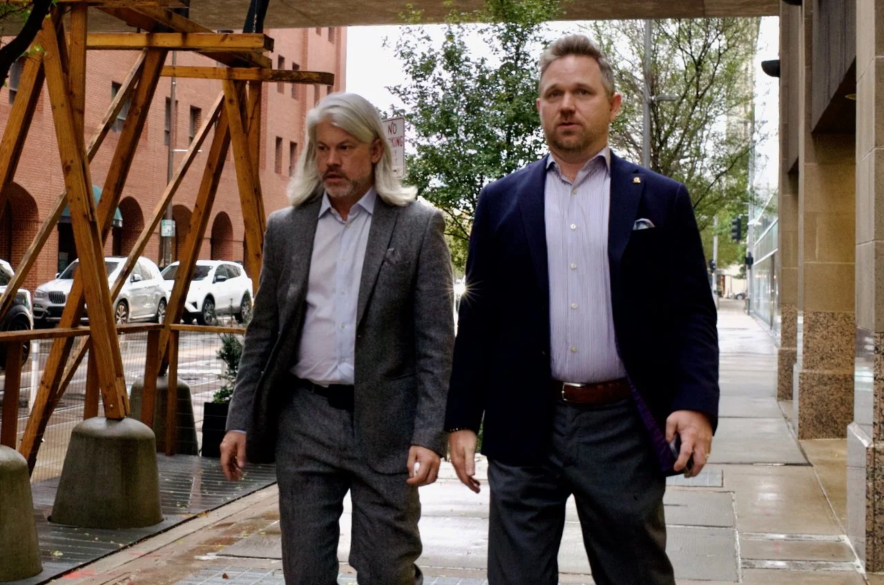 Two men walking on a city sidewalk with buildings and trees in the background.