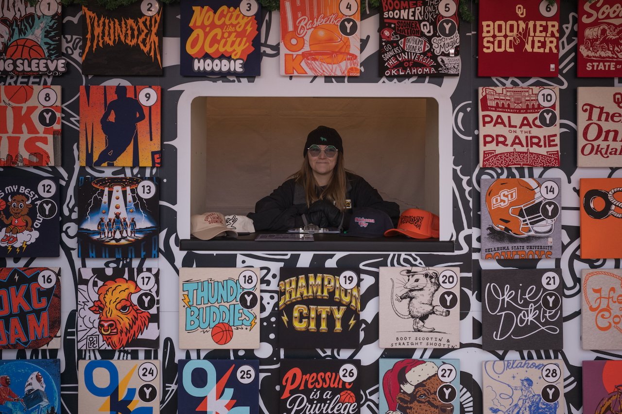 A woman sitting at a kiosk surrounded by Oklahoma-themed and sports-themed graphic posters and banners.