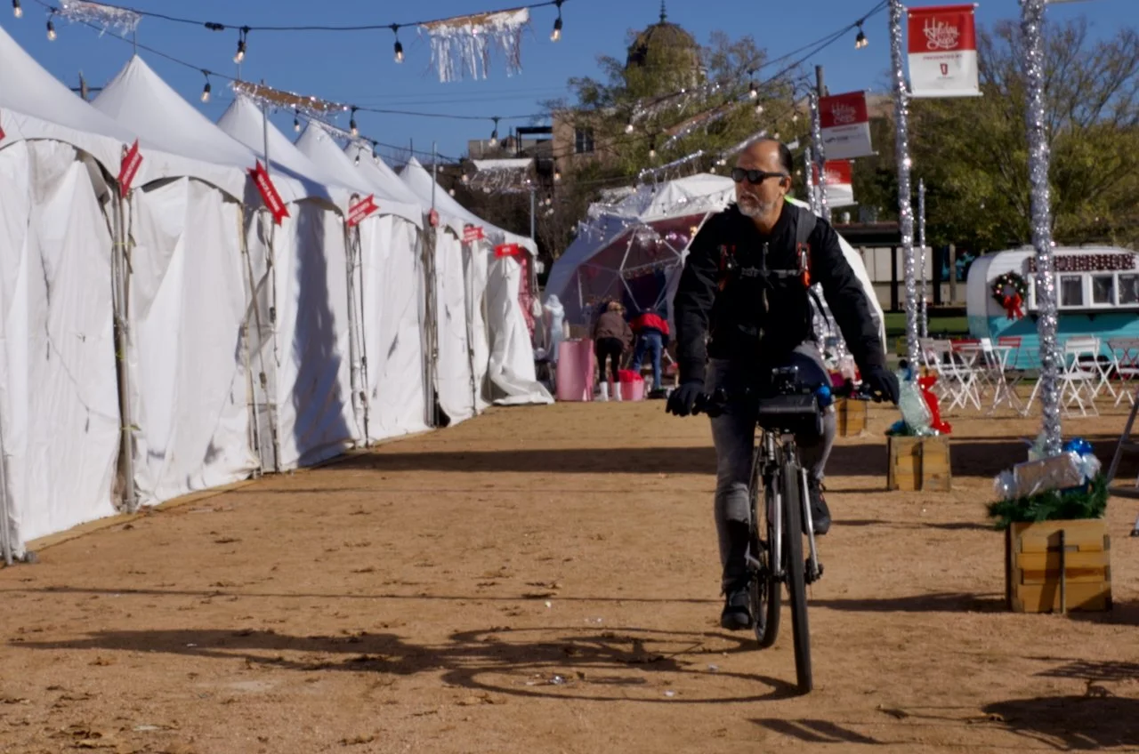 A man riding a bicycle at an outdoor fair or market decorated with string lights and holiday-themed decorations, including tents, umbrellas, and a food truck in the background.