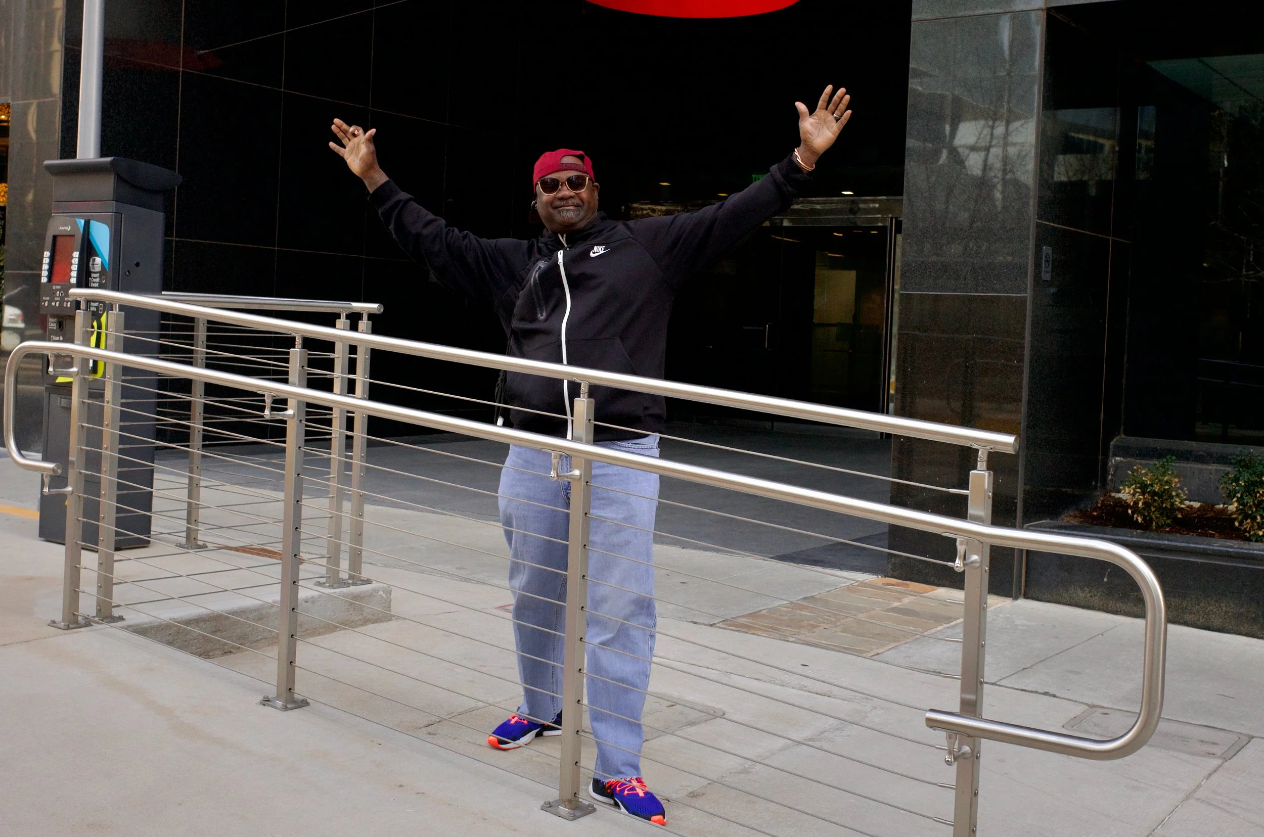 A man standing outside a building with his arms raised and smiling, wearing sunglasses, a red cap backward, a black Nike jacket, blue jeans, and colorful sneakers.