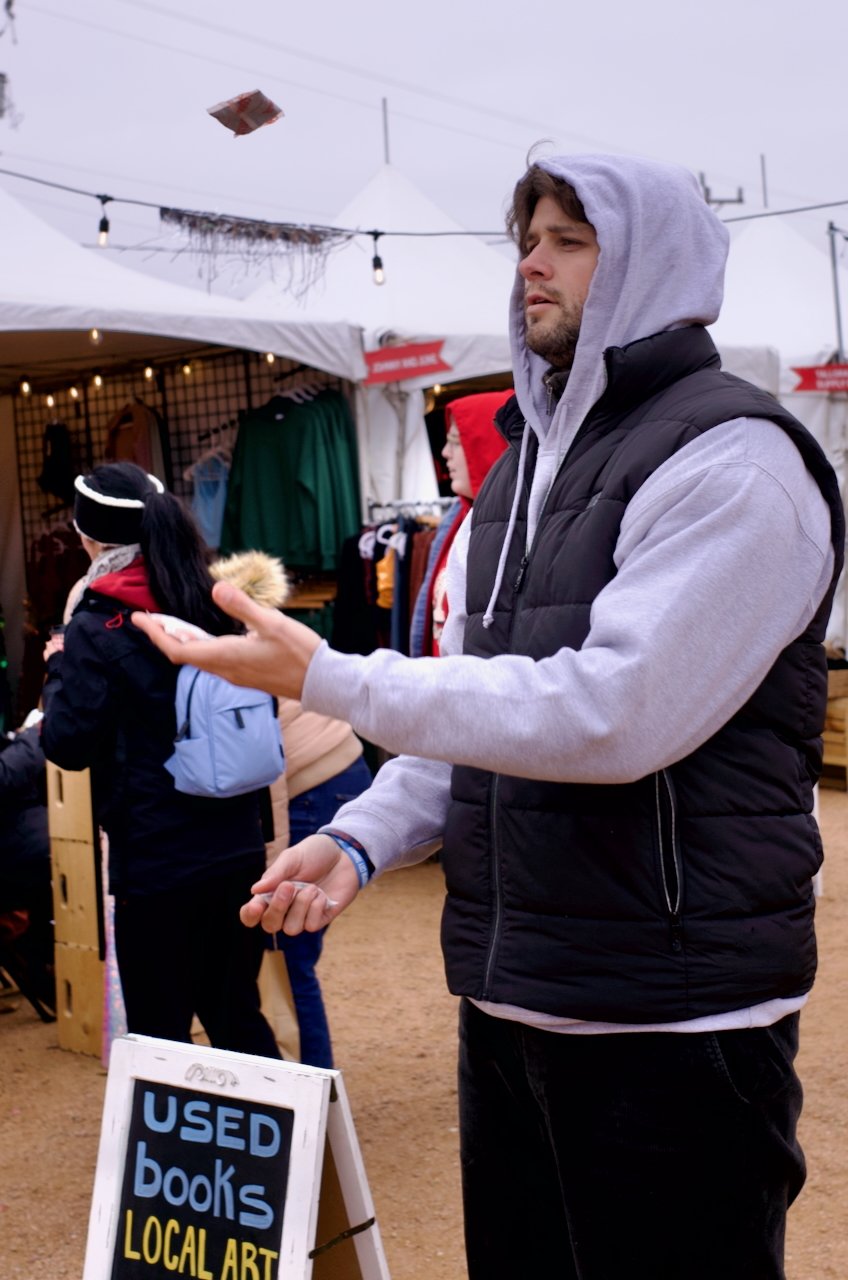 A man wearing a gray hoodie and black vest is tossing a small object at a market stall. The market area has tents, hanging lights, and people shopping, with a sign advertising used books and local art in the foreground.