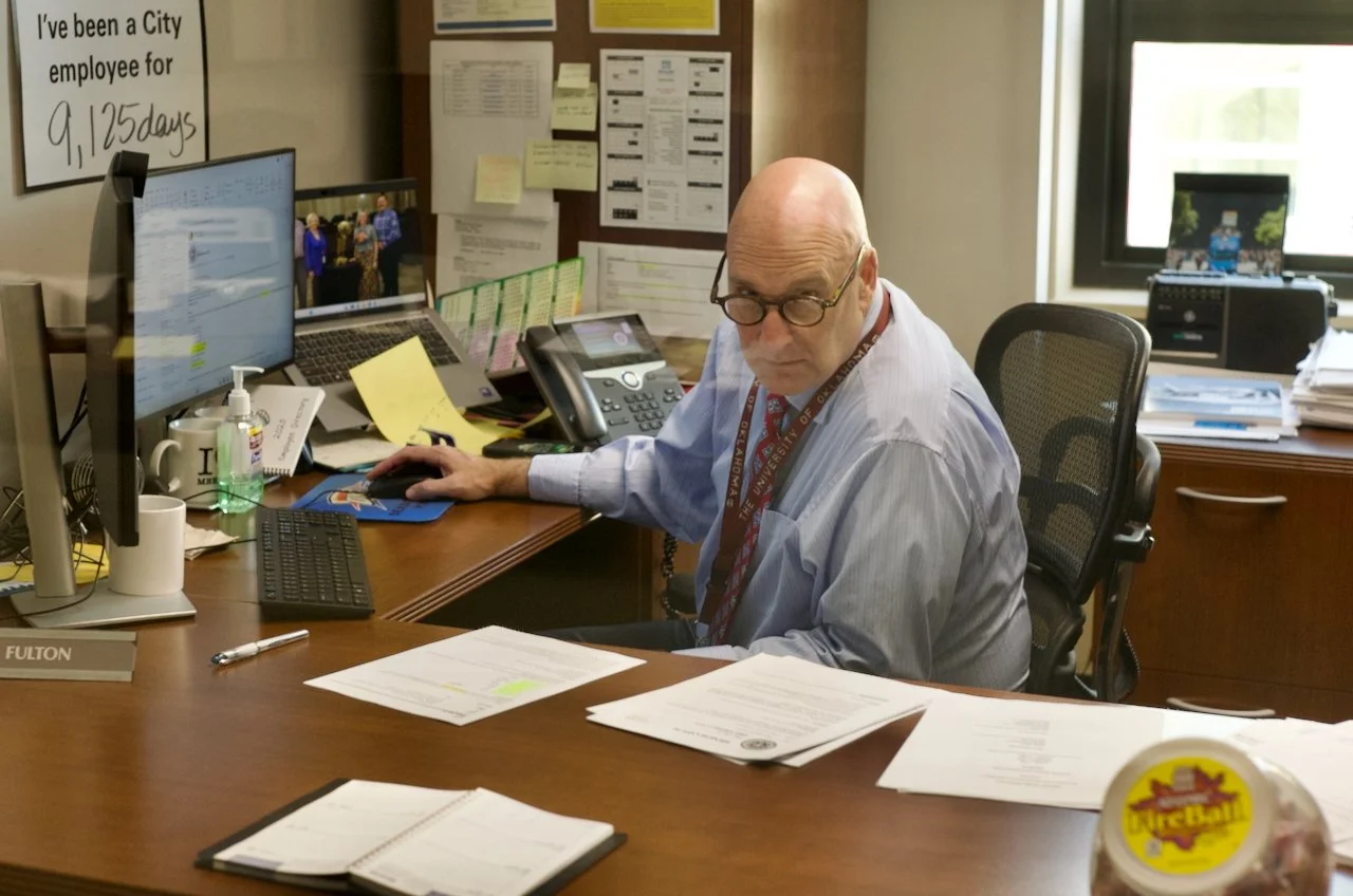 An older man wearing glasses and a light blue shirt seated at a cluttered office desk, working on a computer, with papers, a phone, and office supplies around him. Posters and notes are pinned on the wall behind him.