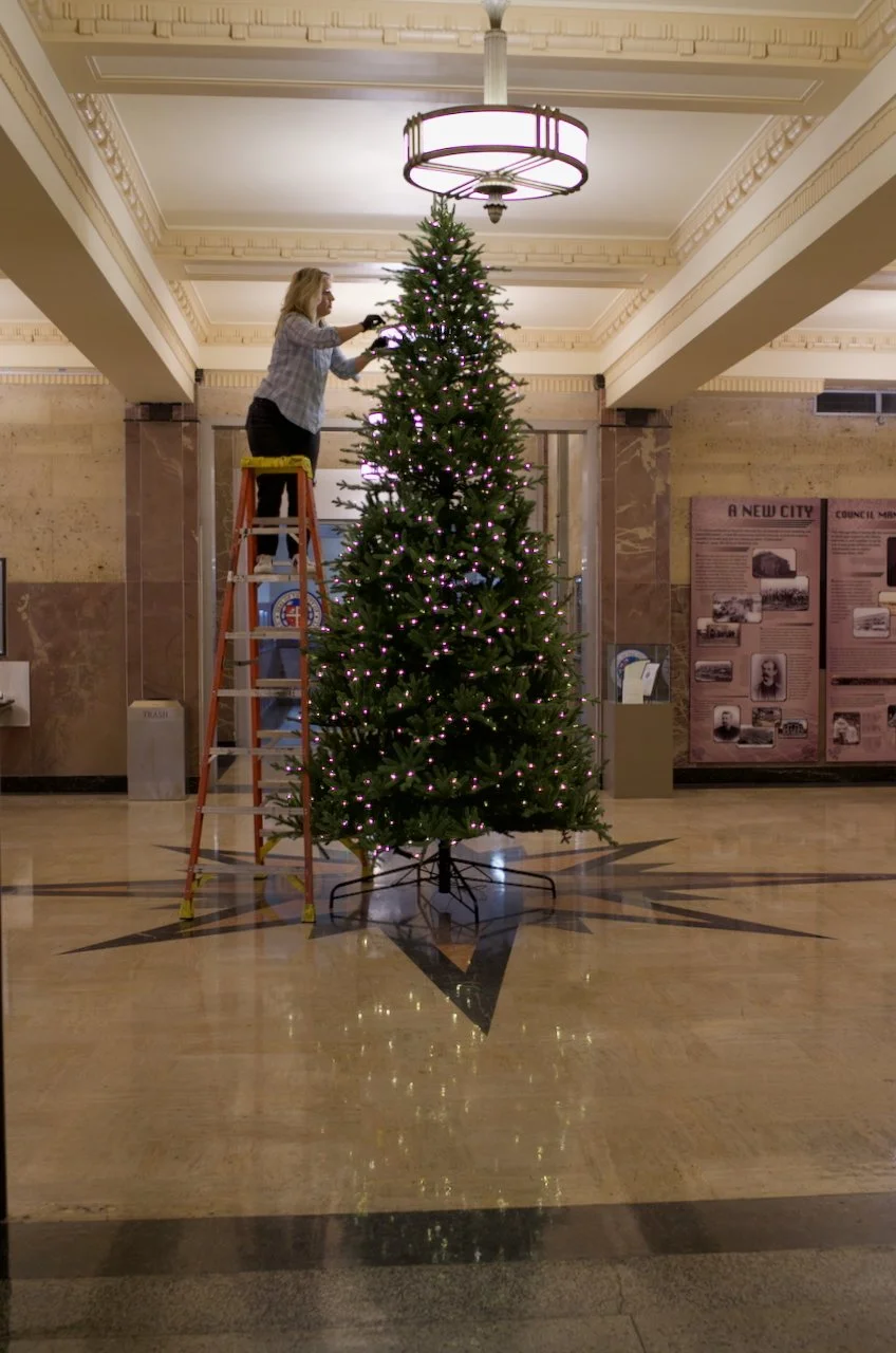 A woman standing on a ladder decorating a tall Christmas tree with pink lights in a spacious lobby with decorative ceiling moldings.