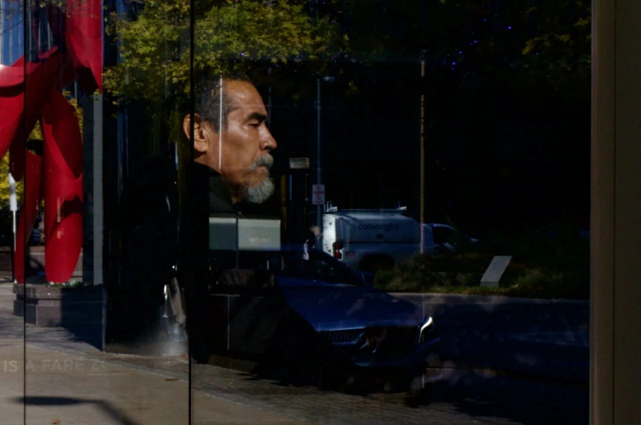 Reflection of a man with gray hair and beard in a glass window, with a crosswalk, parked cars, trees, and a building outside.
