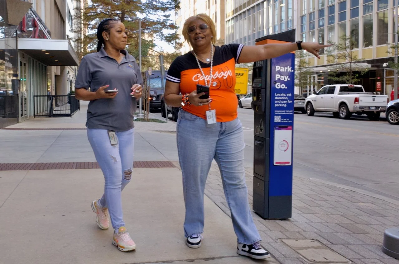 Two women standing on a city sidewalk near parking meters, engaged in conversation; one wearing a Cowboys shirt and pointing, the other in a gray polo and distressed jeans.
