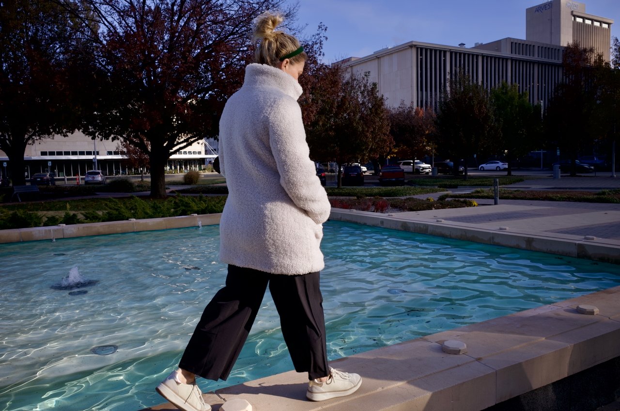 A woman in a white fuzzy coat, black pants, and white sneakers walking along the edge of a water fountain in an urban park during late afternoon or early evening.