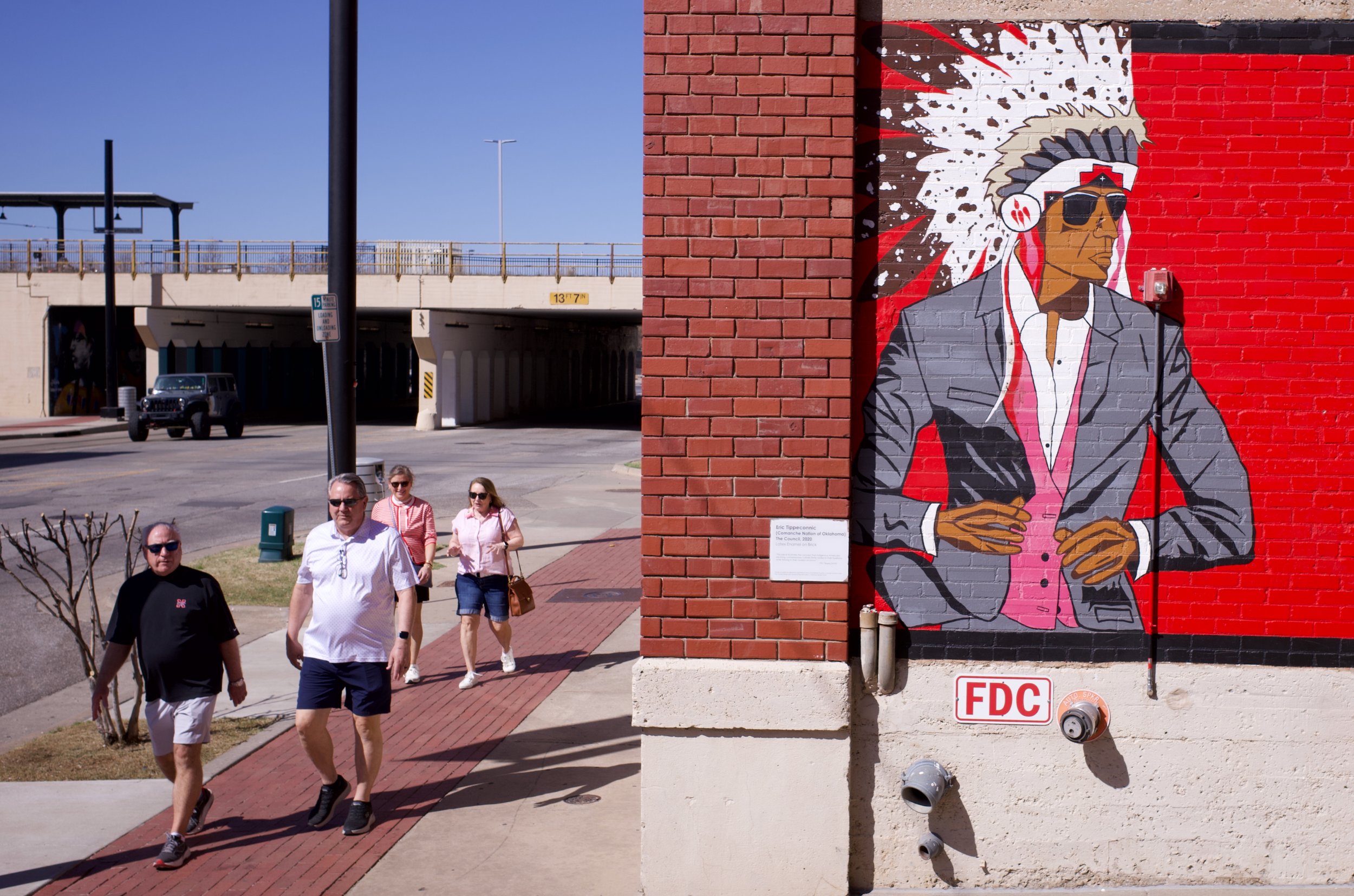 Street scene with four people walking on a sidewalk next to a large mural of a man wearing sunglasses, a grey suit, and a headdress, on a brick wall. The mural has a red background with splatters. There is a parking garage overhead and a dark vehicle
