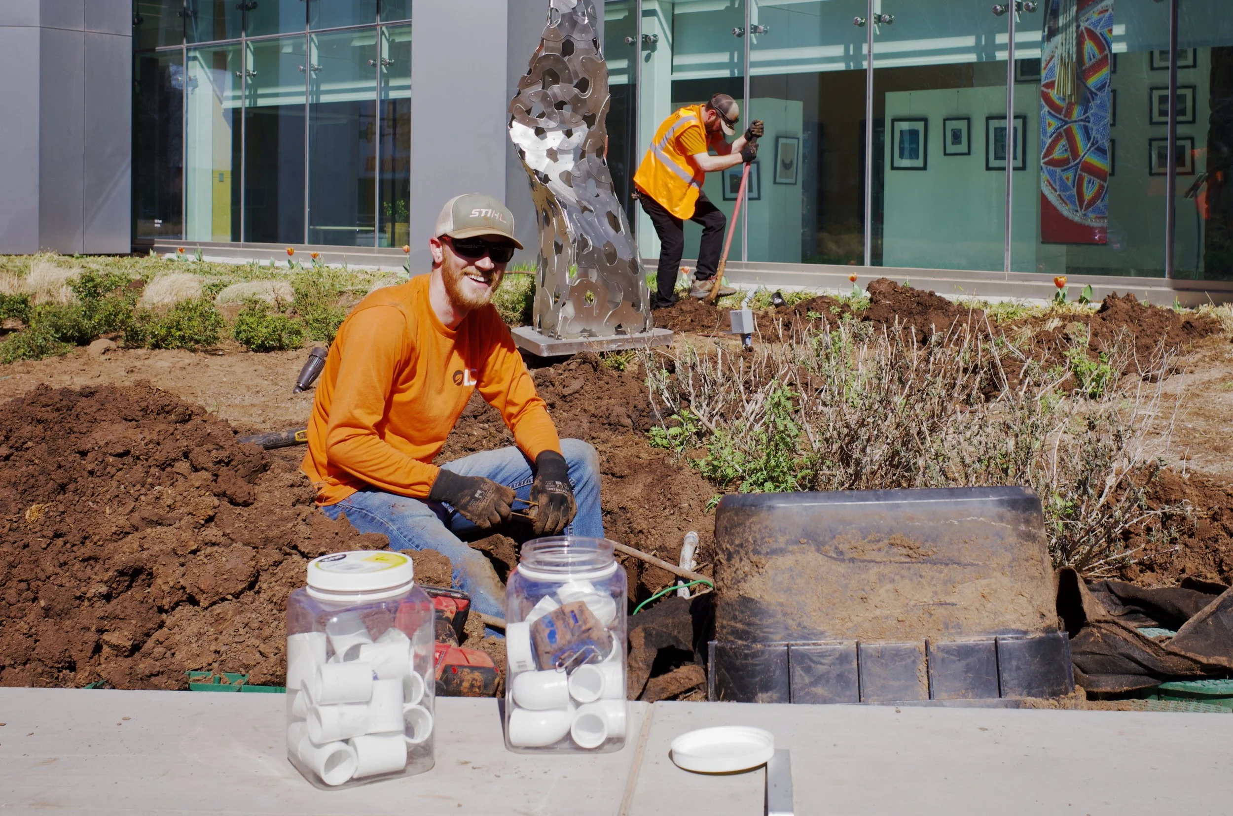 Two workers planting or maintaining plants outside a modern building. One worker is sitting on the ground, smiling at the camera, with gloves and tools. The other worker is standing, wearing safety glasses and a reflective vest, working with soil nea
