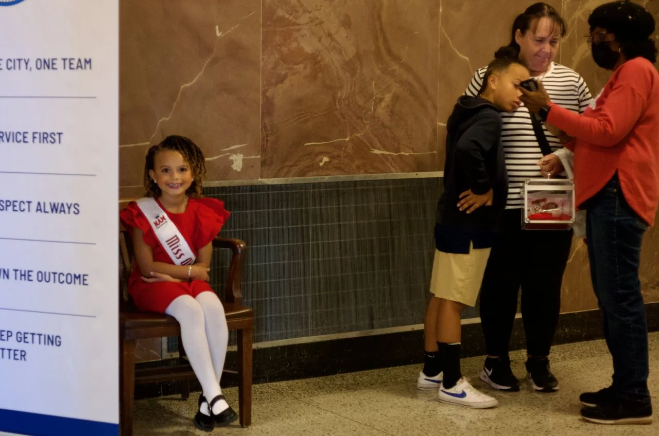 A young girl dressed as Miss Alabama sitting on a wooden bench, smiling at the camera, with curly hair, white tights, and black shoes. To the right, three women and a young boy are standing, with one woman showing something on her phone to the girl a
