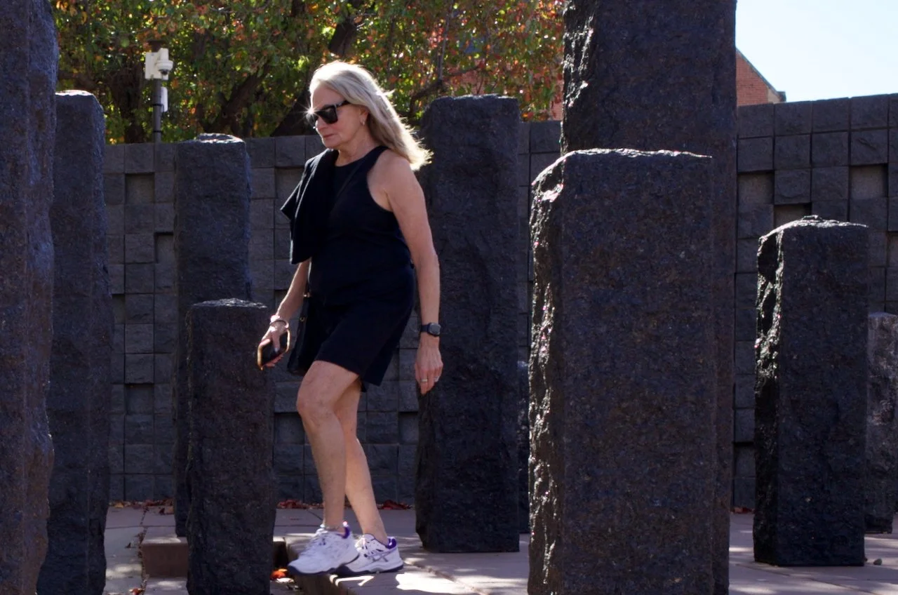 A woman with blonde hair wearing sunglasses, a sleeveless black top, black shorts, and white sneakers walking among tall black stones in an outdoor setting with trees and a wall in the background.