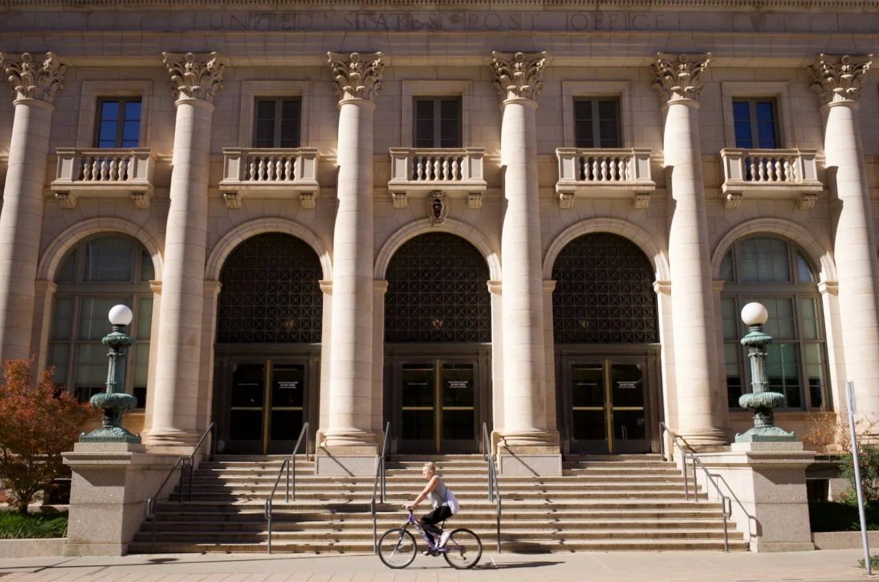 Front entrance of a large stone building with tall columns, arched doors, and balconies above. A woman riding a bicycle is passing by on the sidewalk in front.