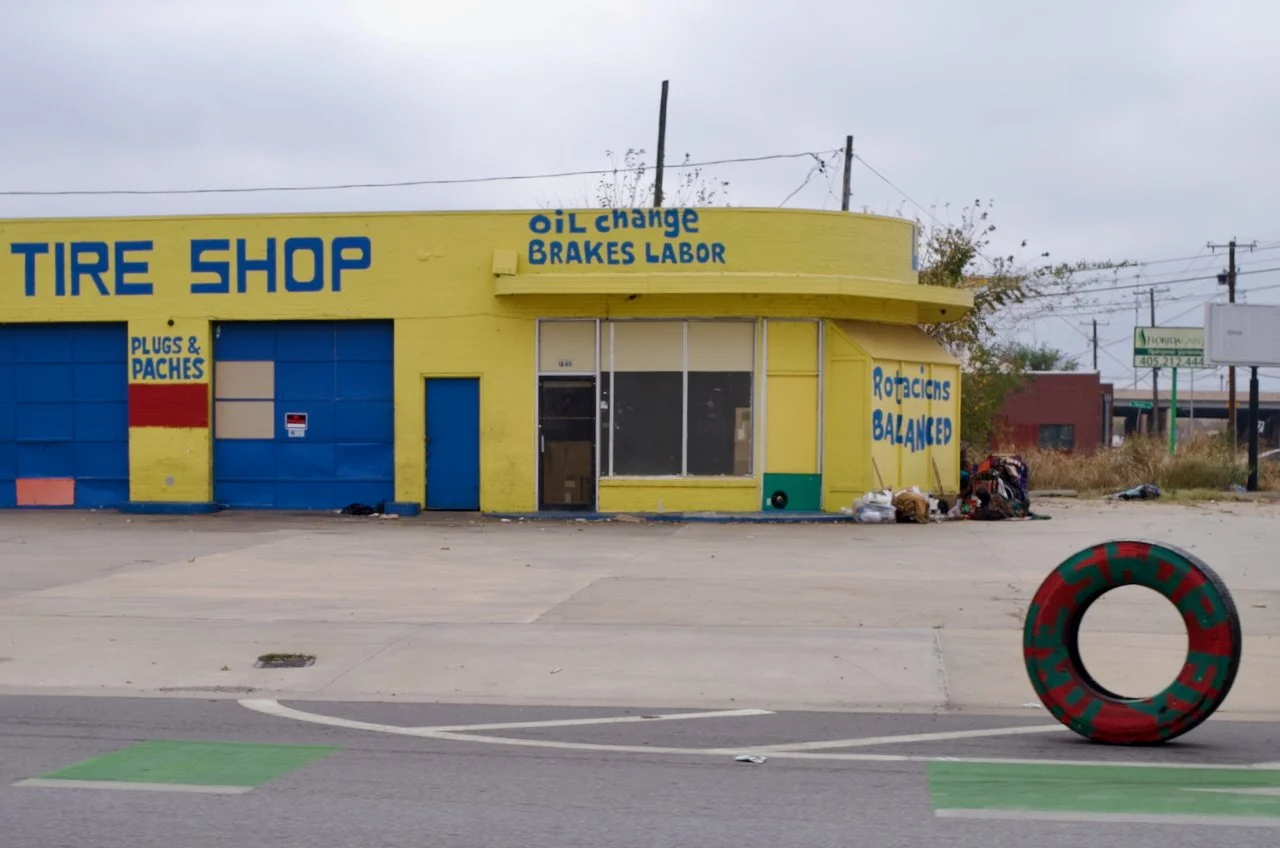 A yellow auto repair shop with blue signage that reads 'Tire Shop', offering services like oil change, brakes, and labor. There are graffiti and bags of trash outside, and a painted tire decoration on the sidewalk.