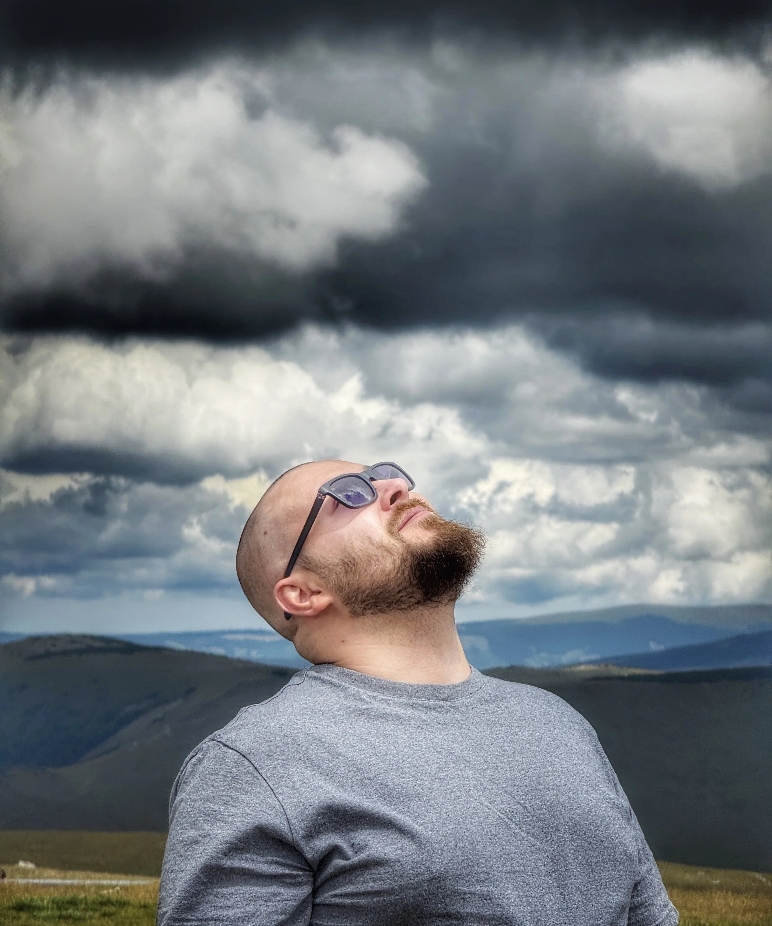 A man with a shaved head, sunglasses, and a beard stands outdoors with his head tilted back, facing the cloudy sky.
