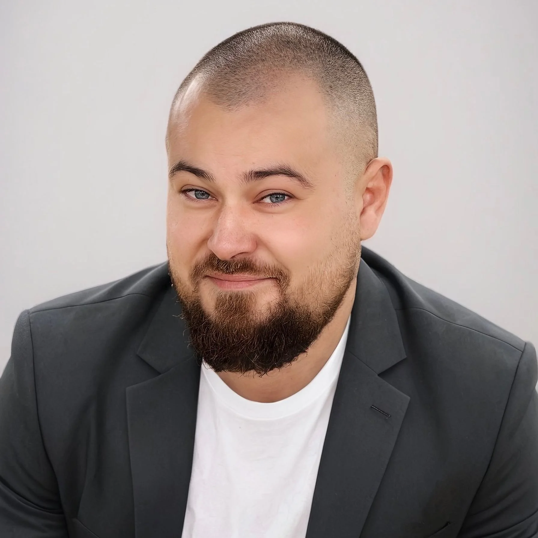 Headshot of a man with a beard and short hair, wearing a dark blazer, smiling slightly, in front of a plain light background.