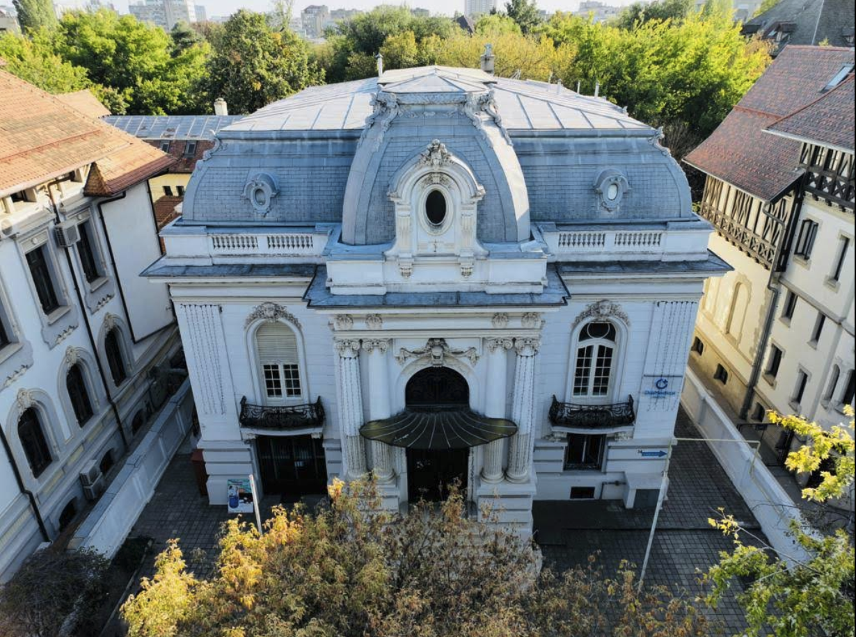 An aerial view of a historic, ornate white building with a curved, gray roof and decorative architectural details, surrounded by other buildings and greenery.