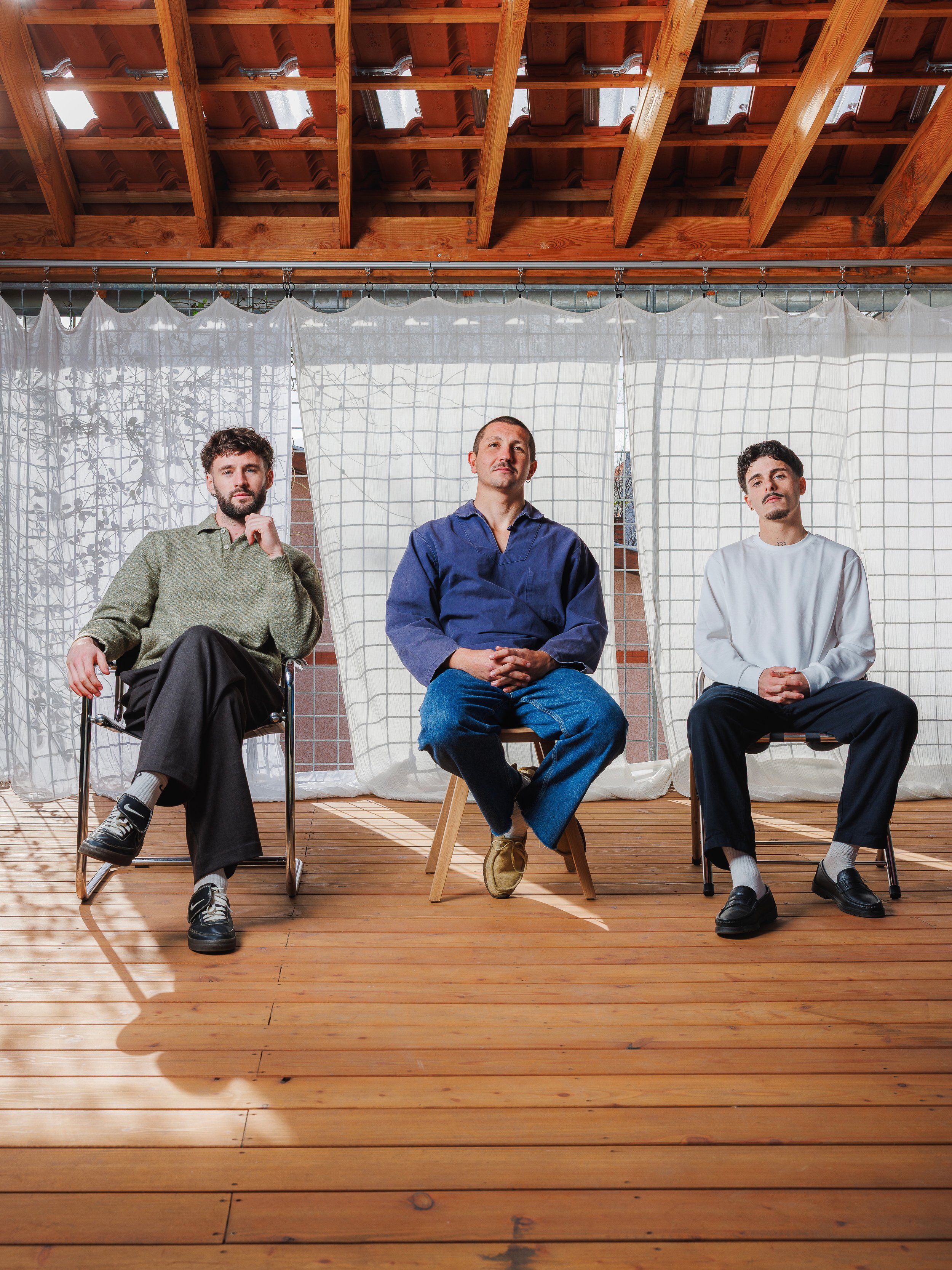 Three men sitting on chairs and stools in front of white curtains in a wooden-ceiling room with sunlight and shadows.