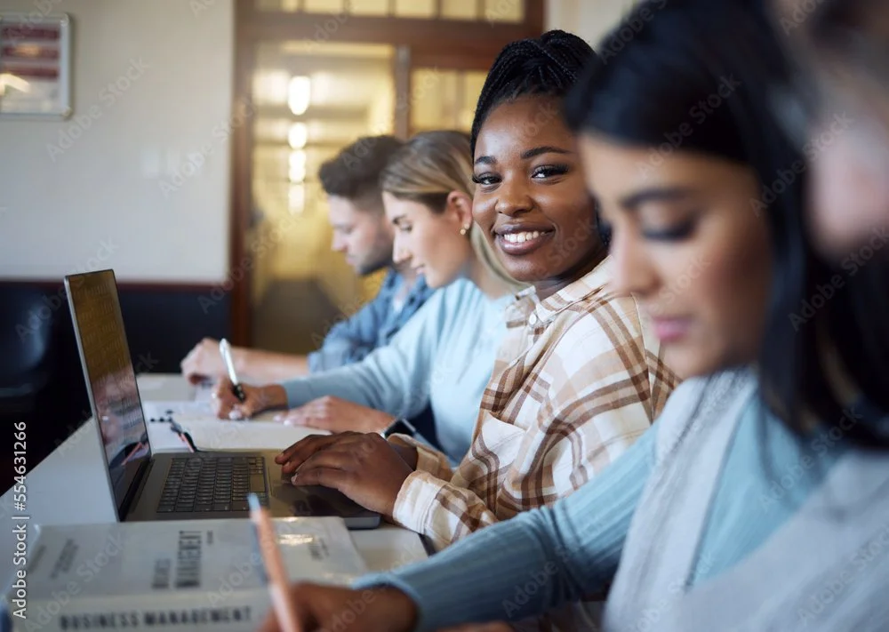 A diverse group of four young adults working on laptops and taking notes in a meeting room.