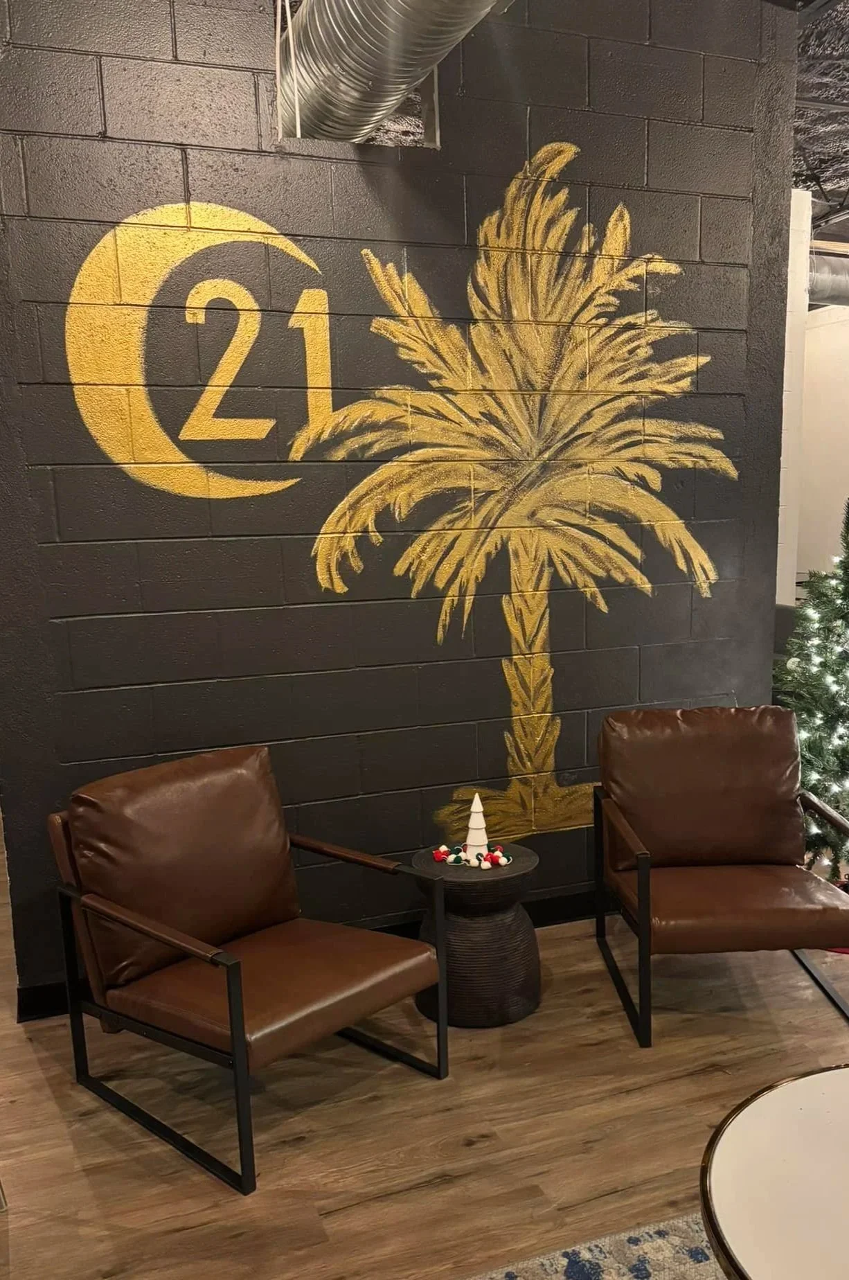 Inside Century 21 803 realty Lexington Office. A wall decorated with a large yellow palm tree and the number 21 inside a yellow crescent moon. In front of the wall, there are two brown leather chairs and a small black side table.
