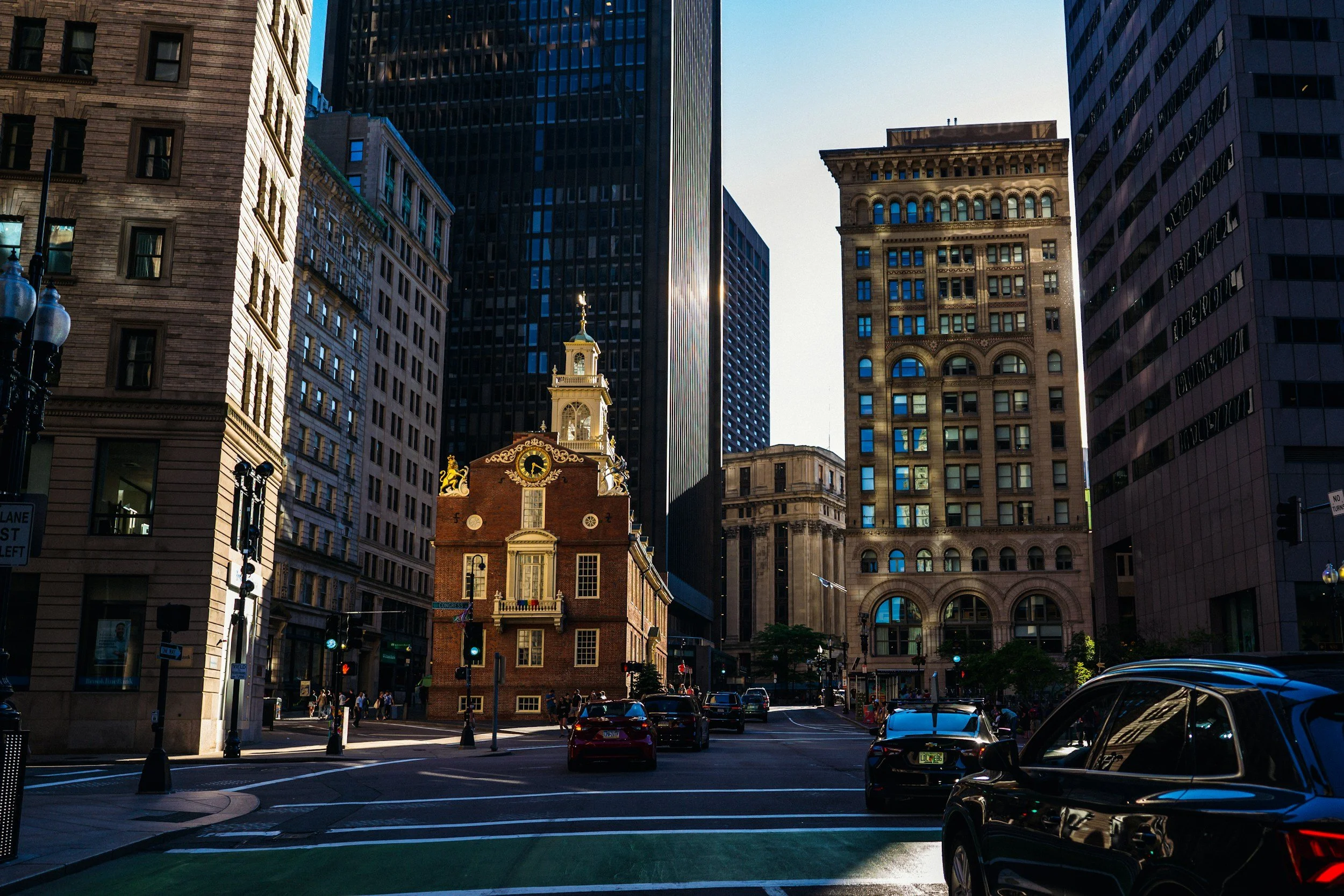 City street scene with tall modern skyscrapers and historic building in the foreground. Cars are parked along the street with a few pedestrians walking on the sidewalk. The sky is clear.