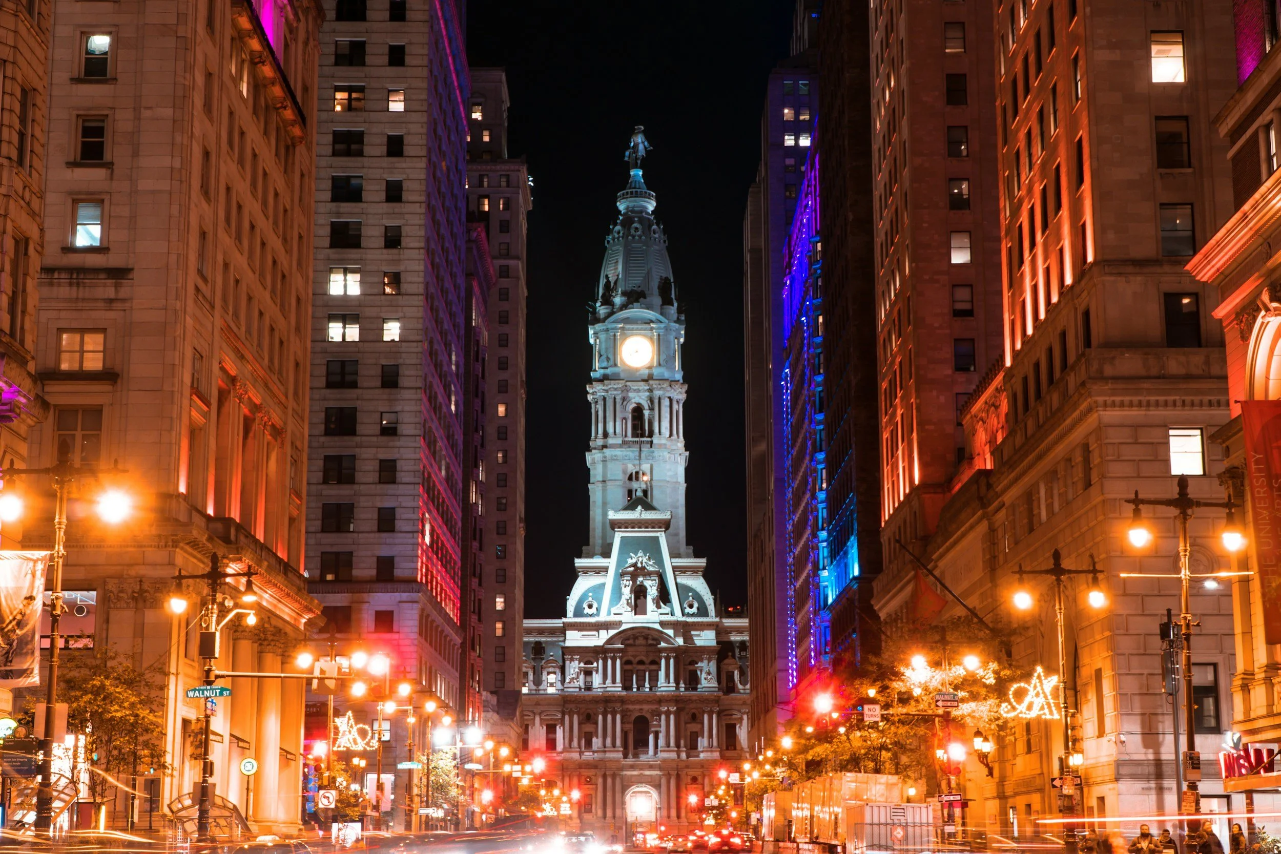 Nighttime view of a city street with tall buildings decorated with colorful lights. A historic clock tower with a statue on top is in the center, illuminated against the dark sky.