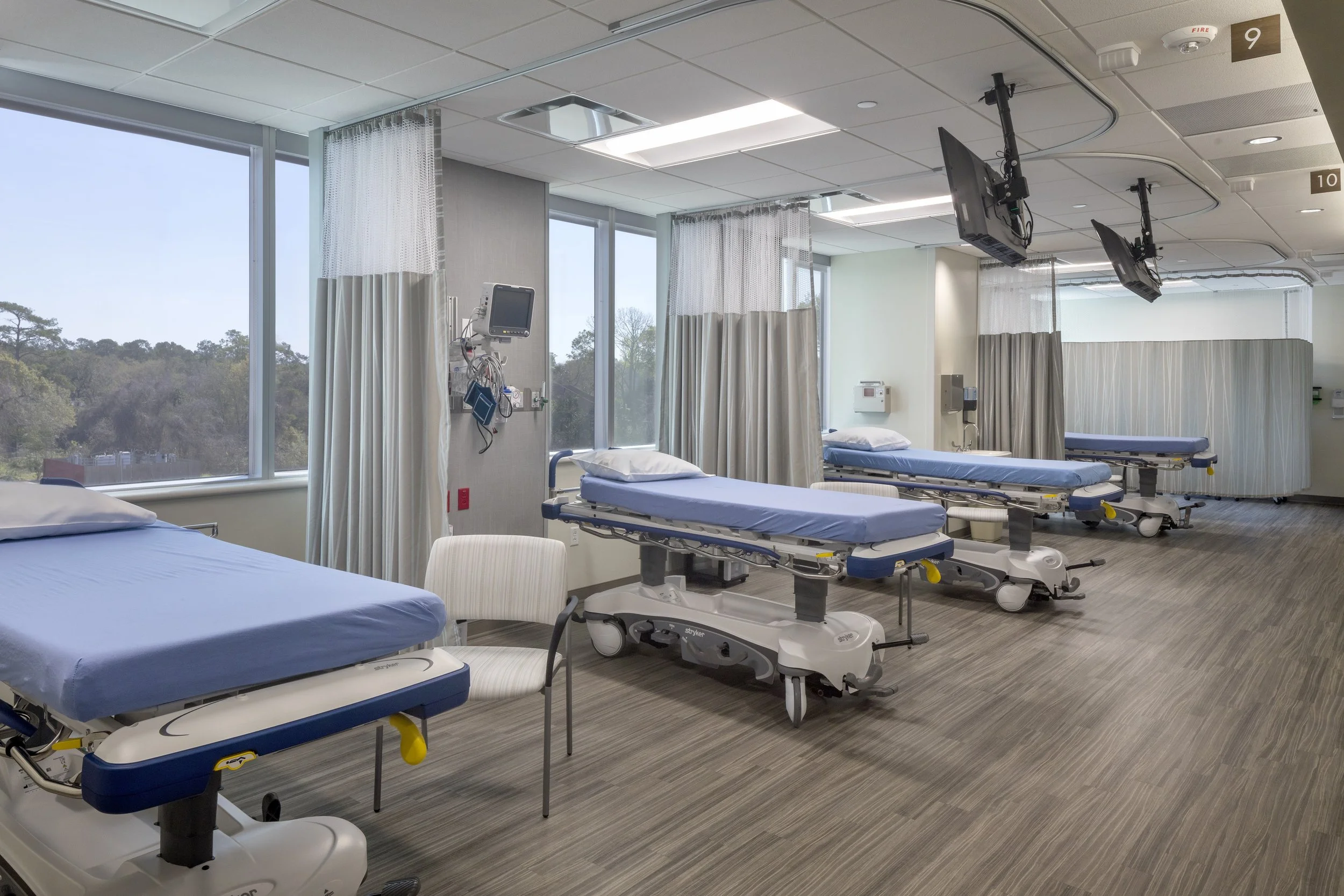 An empty hospital room with four hospital beds, each with a pillow and blue sheet, separated by curtains. Medical equipment, monitors, and a chair are near the beds. Large windows show trees outside, brightening the room with natural light.