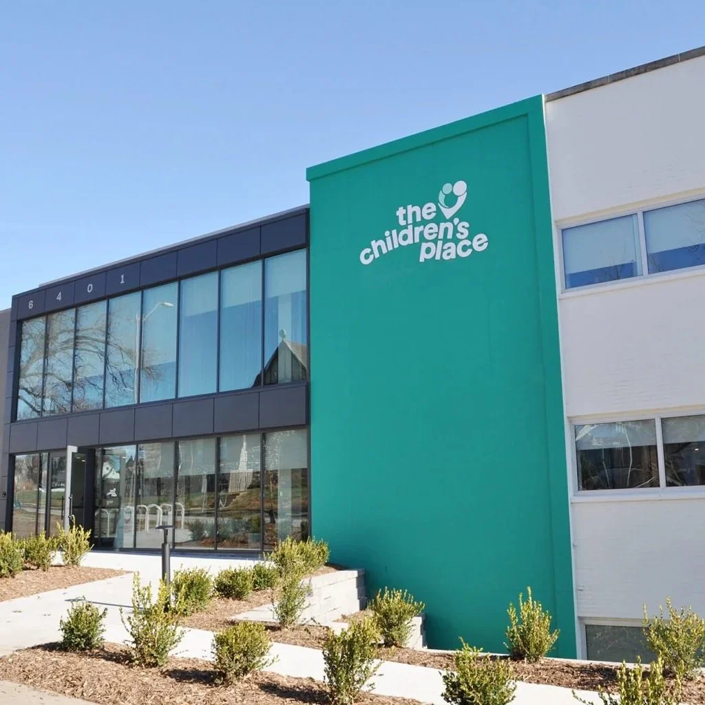 Exterior of a building with a sign that reads "the children's place" on a teal wall, with large glass windows and a white section, under a clear blue sky.