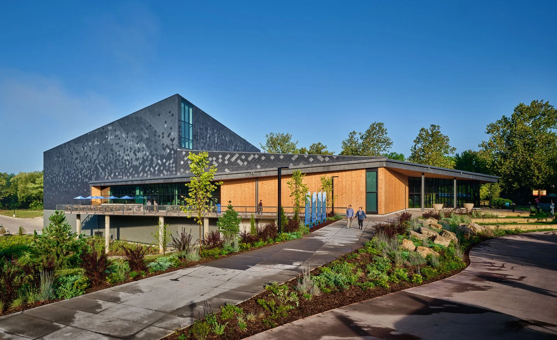 Modern building with wood and dark metal exterior, surrounded by landscaped greenery and a sidewalk, with people walking.