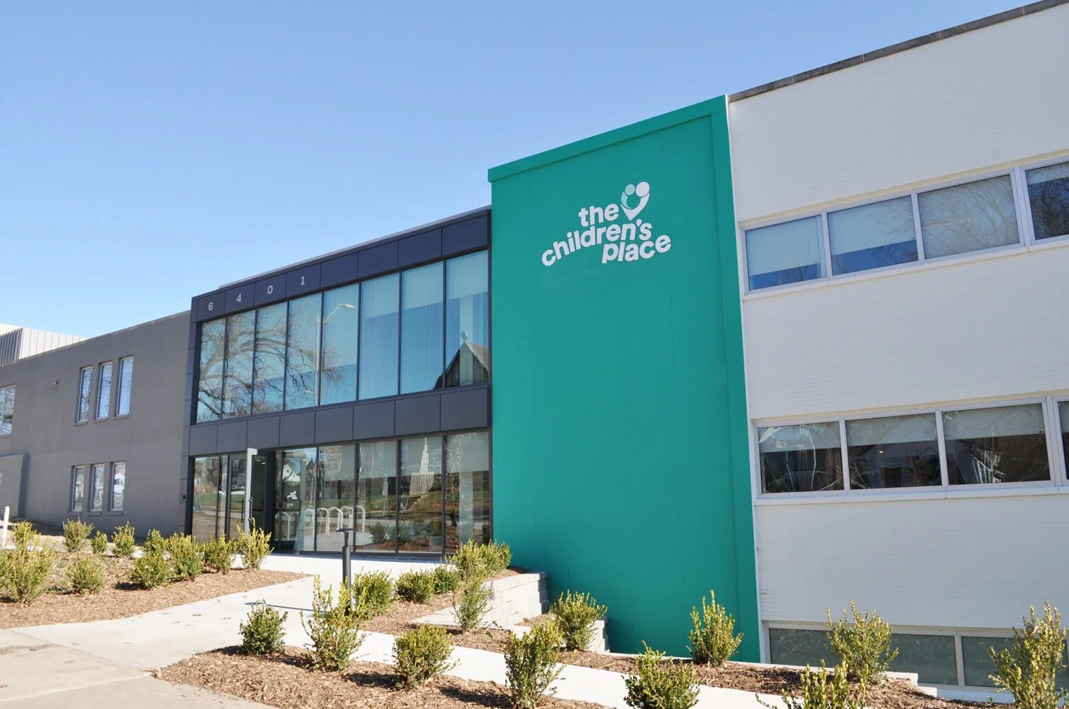 Exterior of a modern building with a sign that reads 'the children's place' on a teal vertical section of the facade, with large windows and landscaped bushes in front, under a clear blue sky.