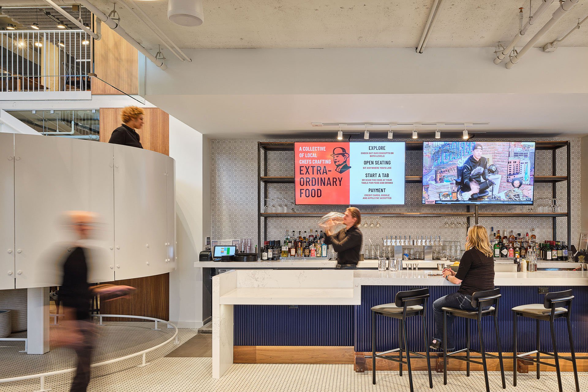 A modern bar with three women, one bartending, one sitting, and one walking past, inside a restaurant with a digital menu on the wall and a large screen showing a person in the background.