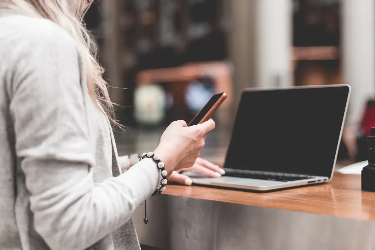 Person in a grey sweater using a smartphone at a wooden table with an open laptop and a camera to the right.