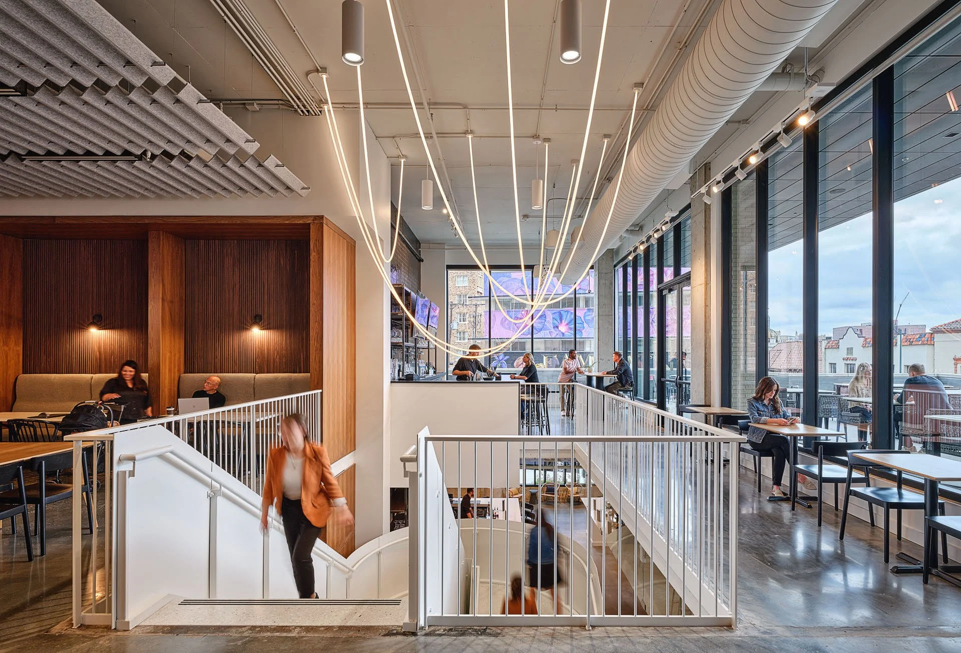Modern cafe or restaurant with large floor-to-ceiling windows, seating on the upper and lower levels, and decorative lights on the ceiling. Customers are seated at tables and at the bar, while a woman walks down the stairs.