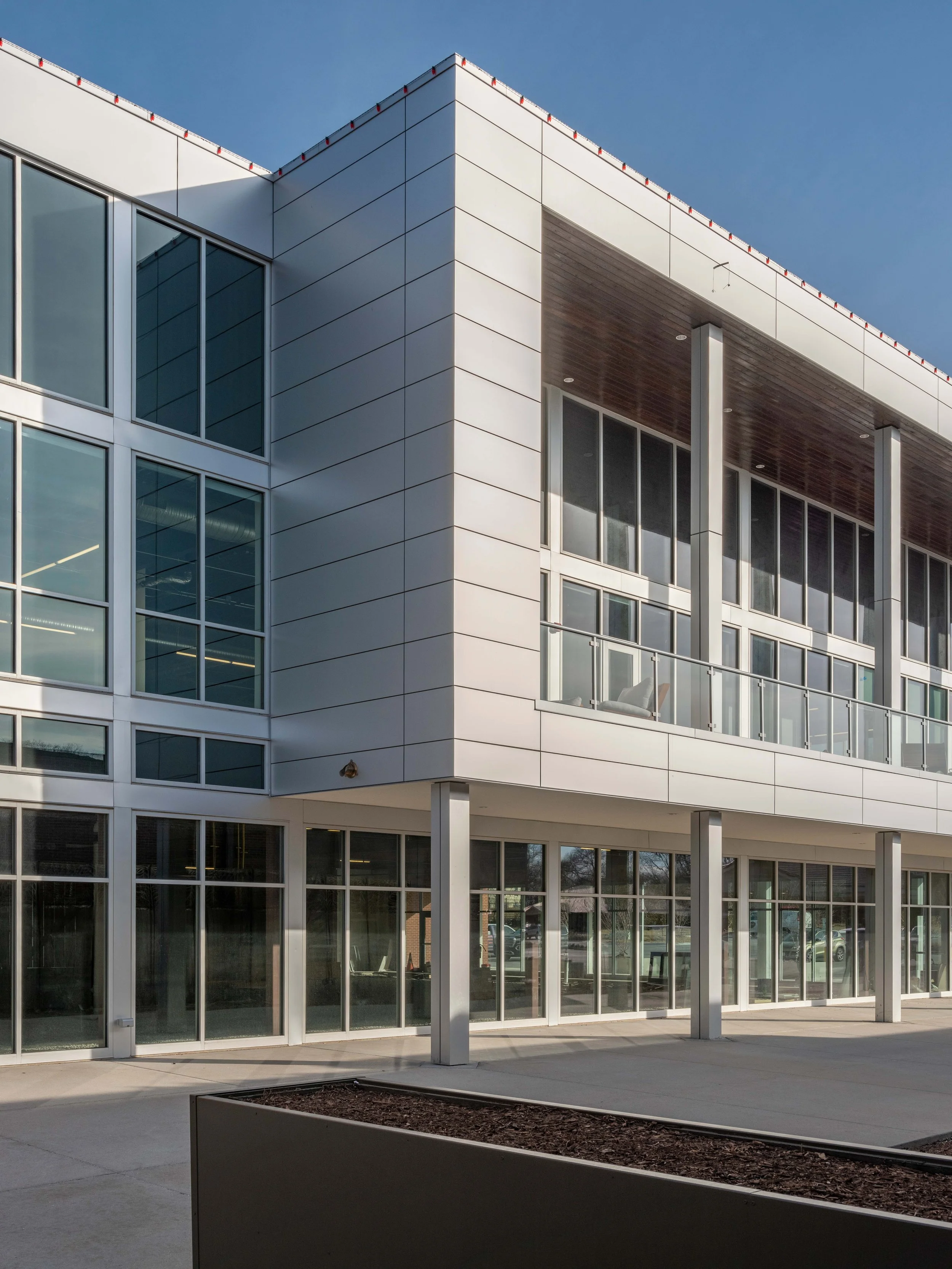 Modern multi-story office building with glass windows and metal paneling, featuring a balcony with glass railing and a small outdoor area in front.