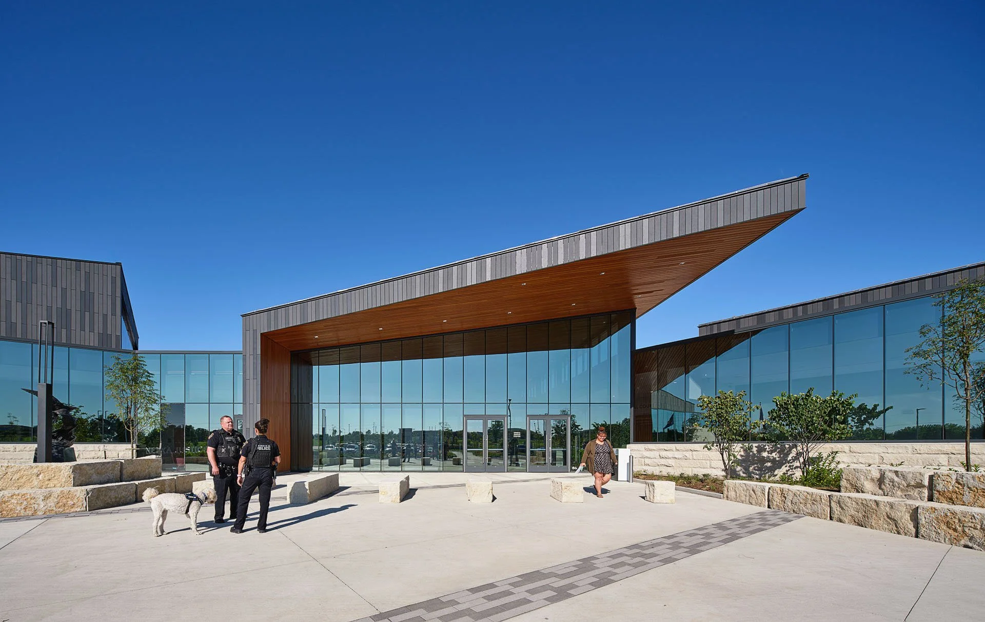 Modern building with large glass windows and a unique angled roof, with three people and a dog standing outside on a sunny day.