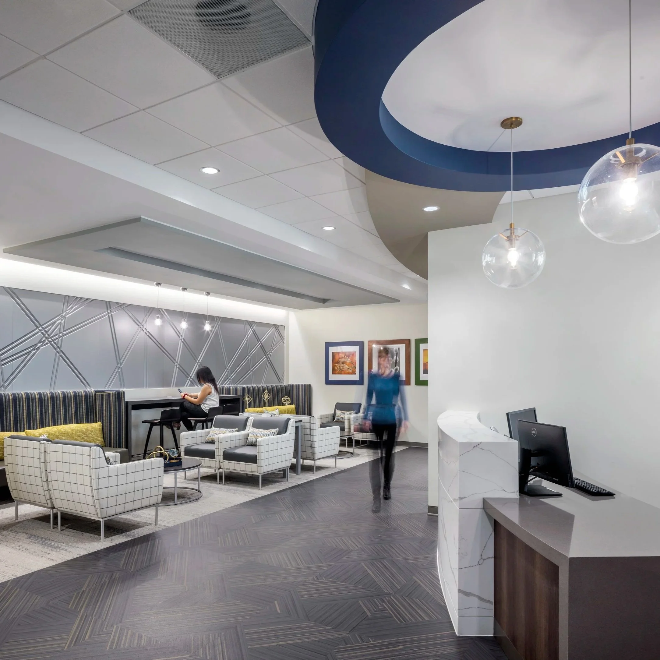 Modern hotel lobby with stylish seating area, a reception desk with a computer, and abstract wall art. Two people are present, one sitting and another walking, with one appearing blurred.