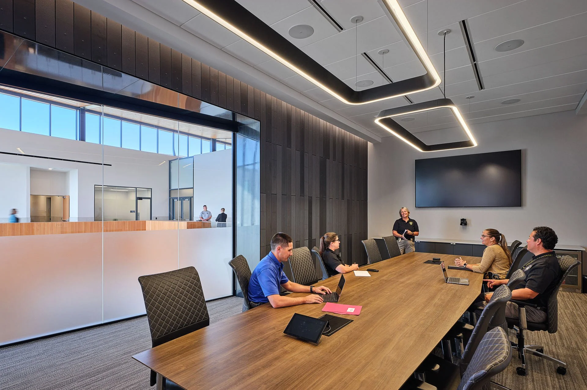 A meeting room with five people sitting at a long wooden table, with one person standing and presenting. Laptops and notepads are on the table, and a large flat screen is mounted on the wall. The room has modern lighting and large windows with a view of an adjacent room.