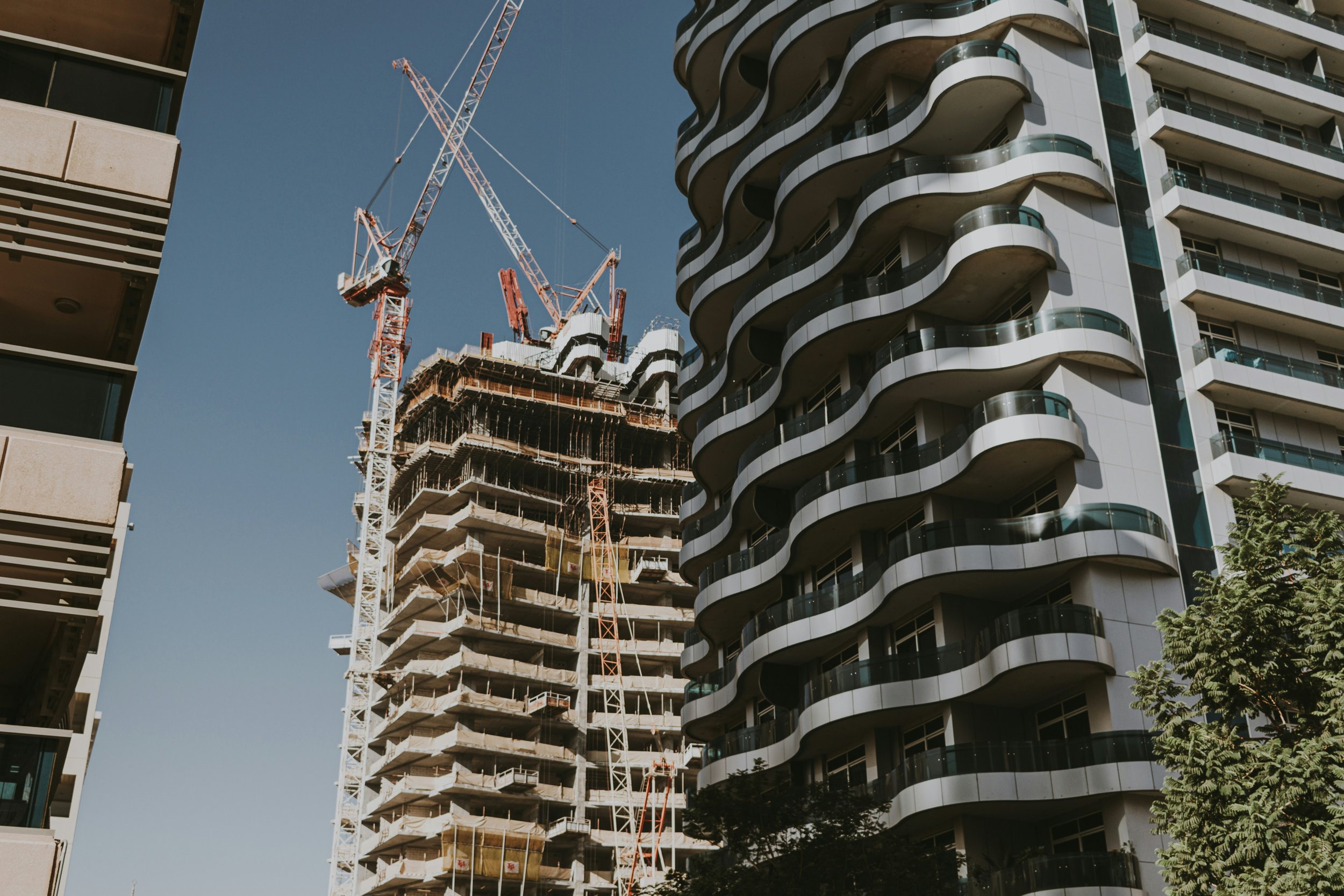 Construction site with a high-rise building under development, cranes, and a completed modern building with curved balconies, under a clear blue sky.