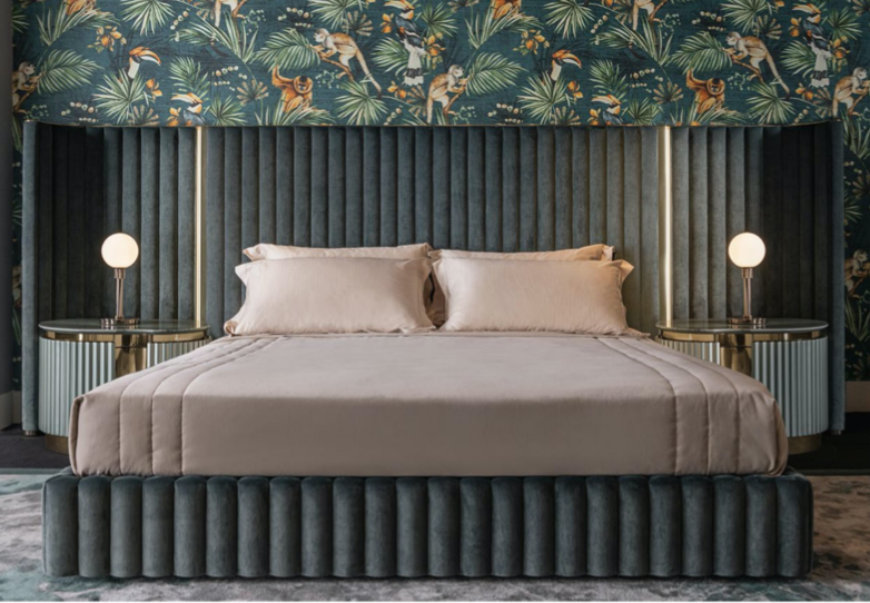 A modern hotel bedroom with a velvet-upholstered bed, beige bedding, two round bedside tables with spherical lamps, and tropical wallpaper featuring leaves and animals.